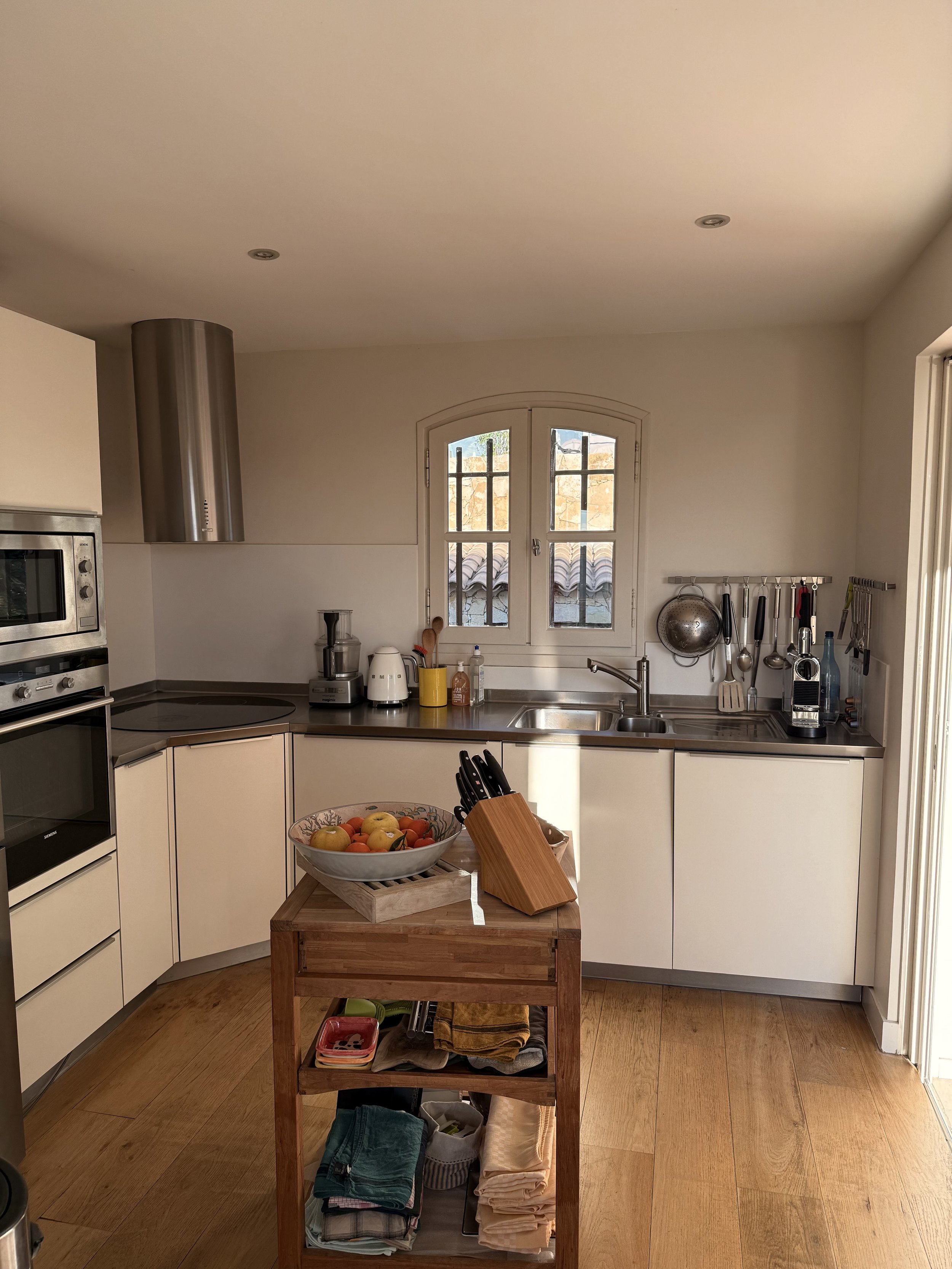 Cuisine moderne avec un comptoir en bois, une fenêtre derrière le lavabo, des ustensiles de cuisine suspendus, un bol de fruits sur un îlot en bois, et des appareils électroménagers en acier inoxydable.