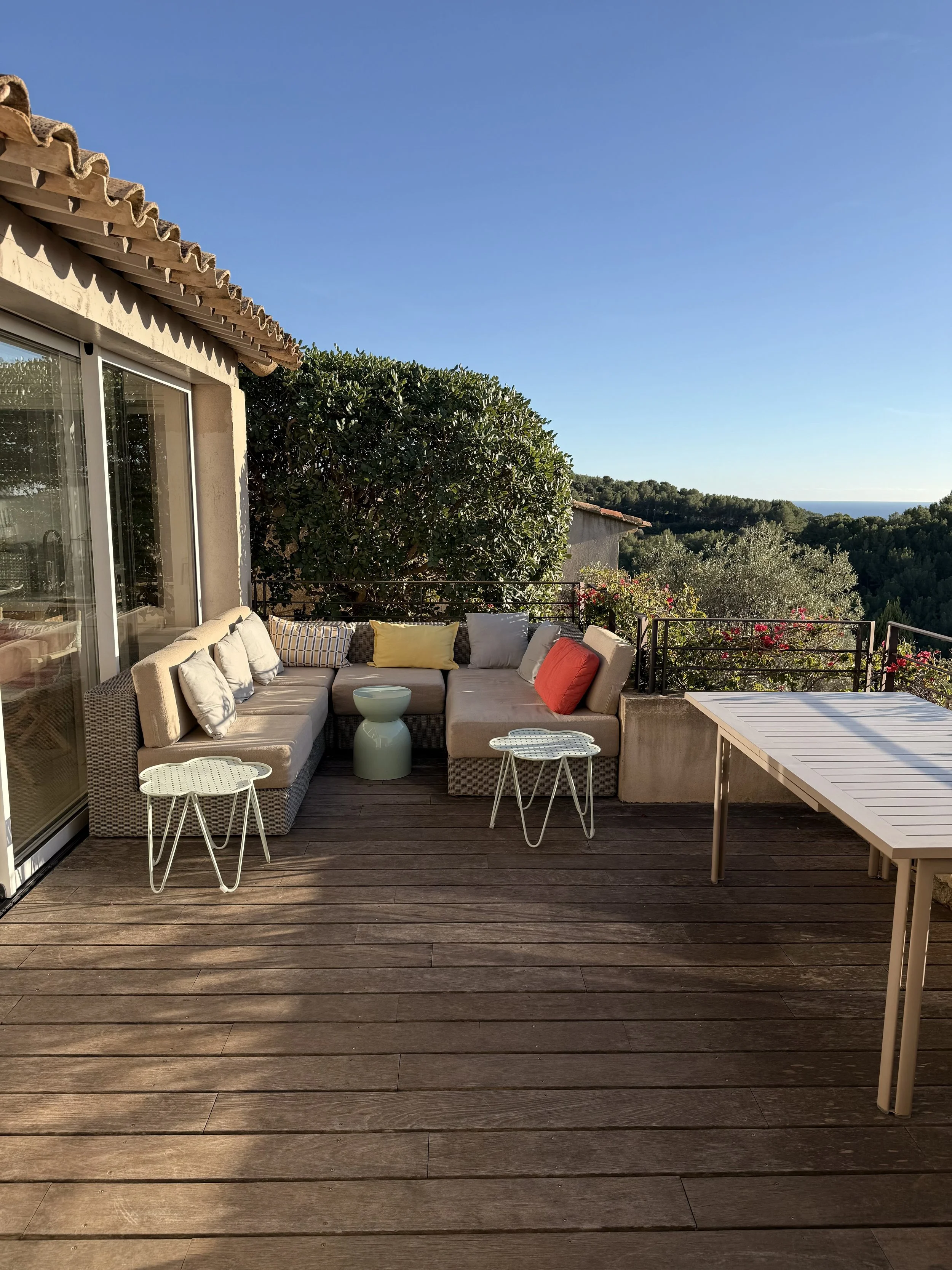 Vue d'une terrasse en bois avec un coin salon entre lui, des tables d'appoint, un grand arbre en arrière-plan et une vue vers la campagne avec des arbres et le ciel clair.