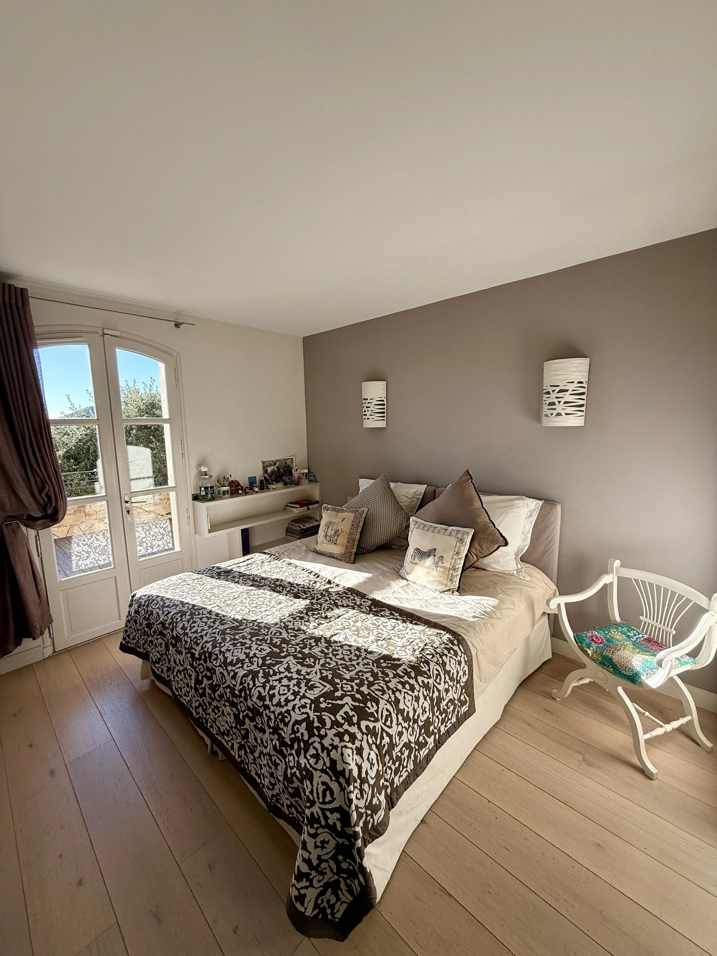 Chambre avec un lit, des coussins, une chaise blanche, et des fenêtres laissant entrer la lumière naturelle.