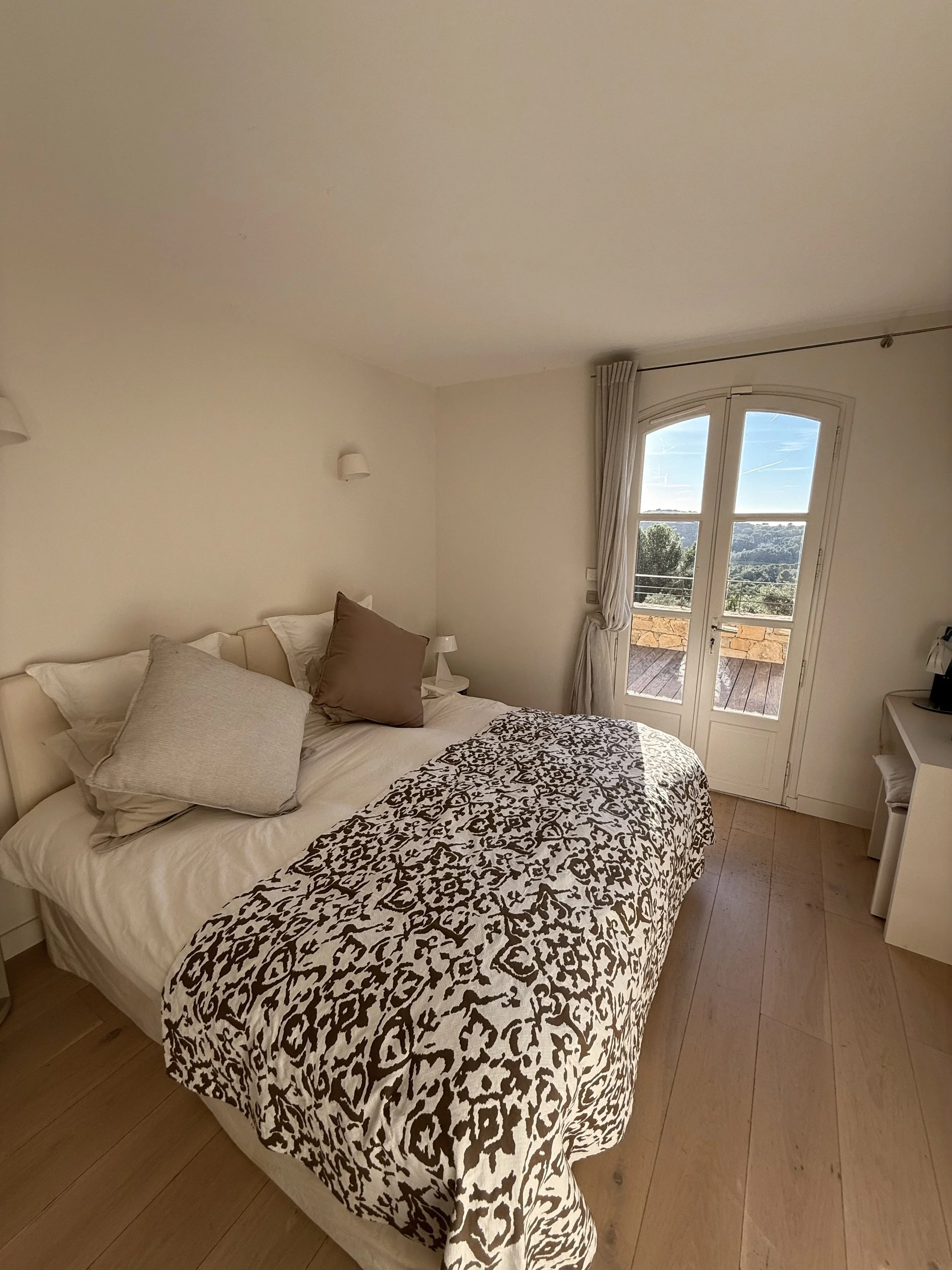 Chambre avec lit, coussins et couverture à motif, porte-fenêtre donnant sur un balcon avec vue sur la nature.