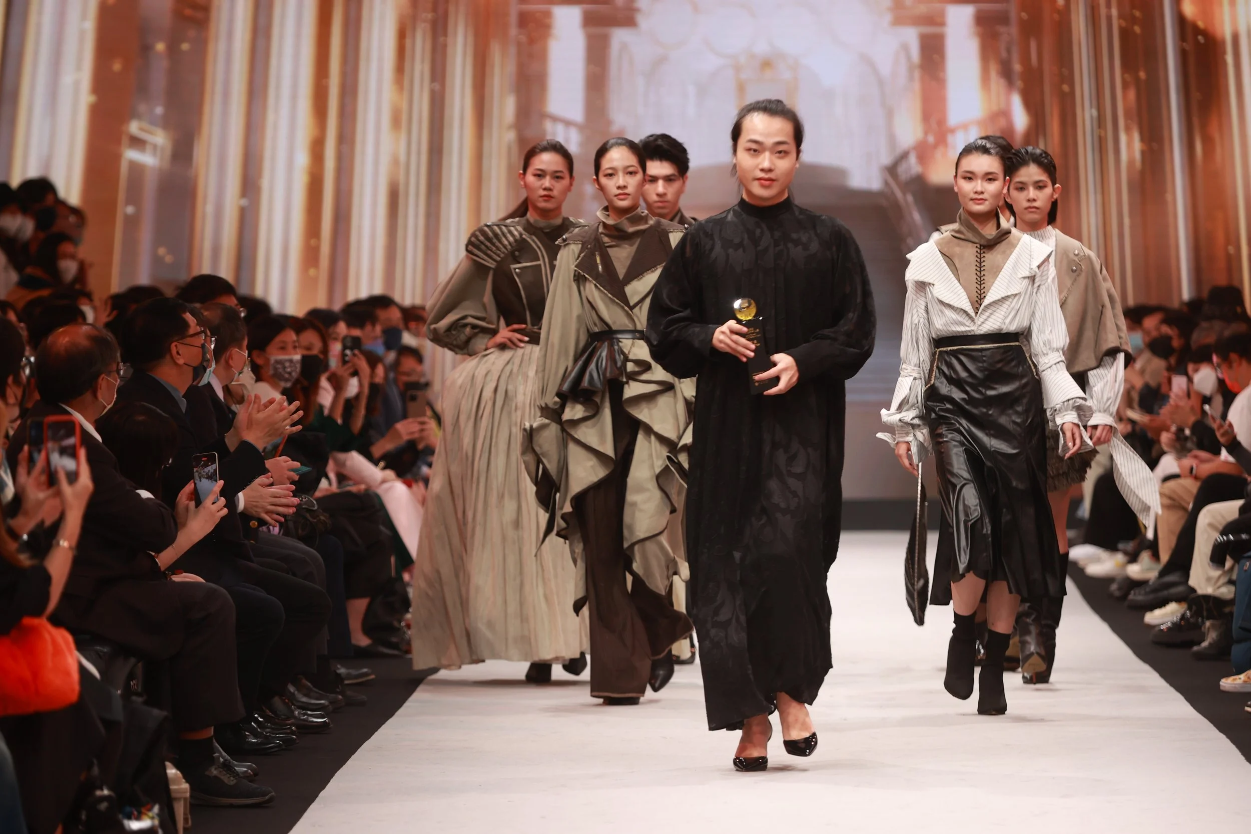 Group of models walking on a fashion runway with an audience seated on both sides.