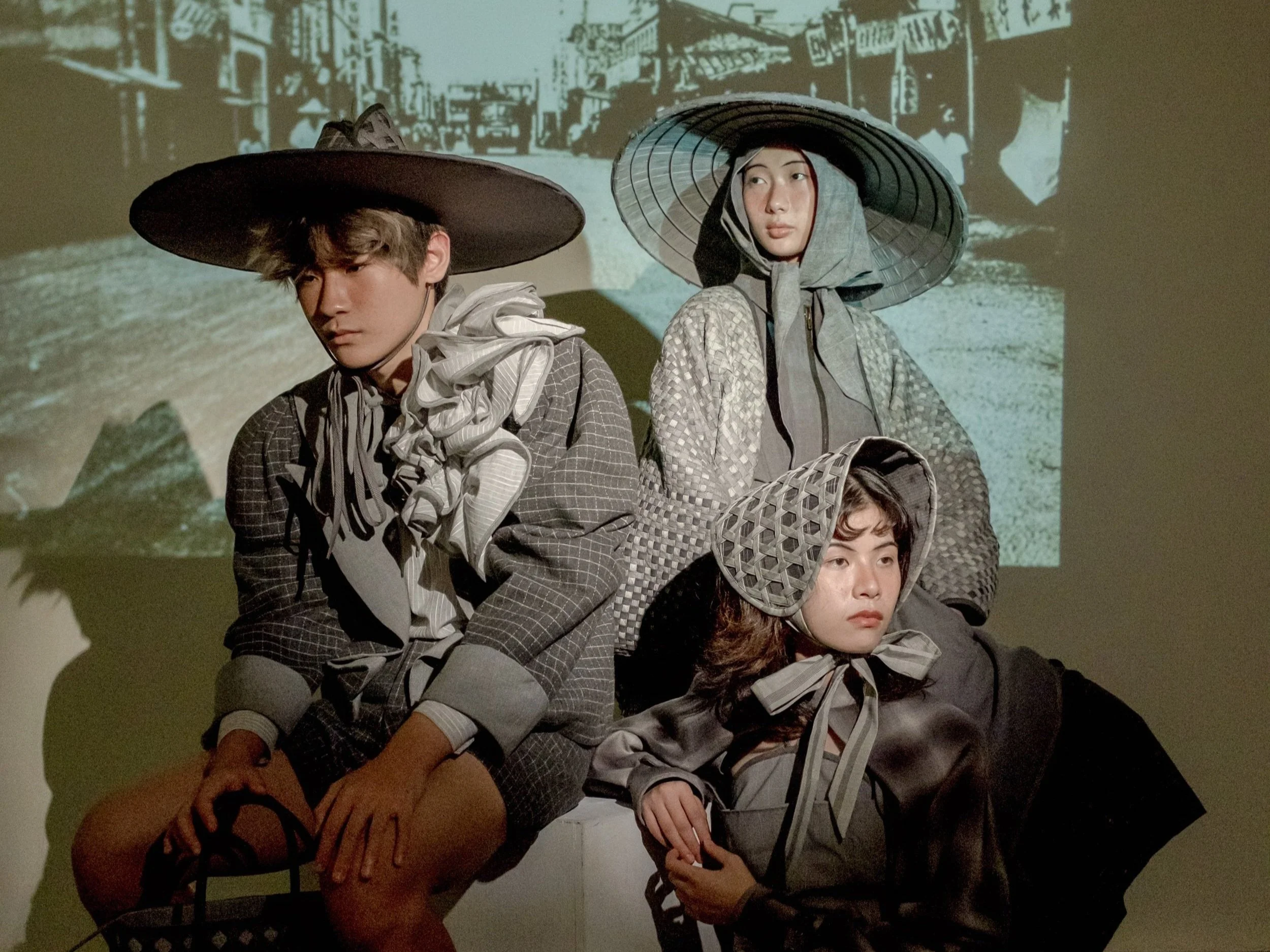 Four individuals dressed in traditional East Asian clothing and wide-brimmed straw hats, sitting in front of a projected black-and-white cityscape background.