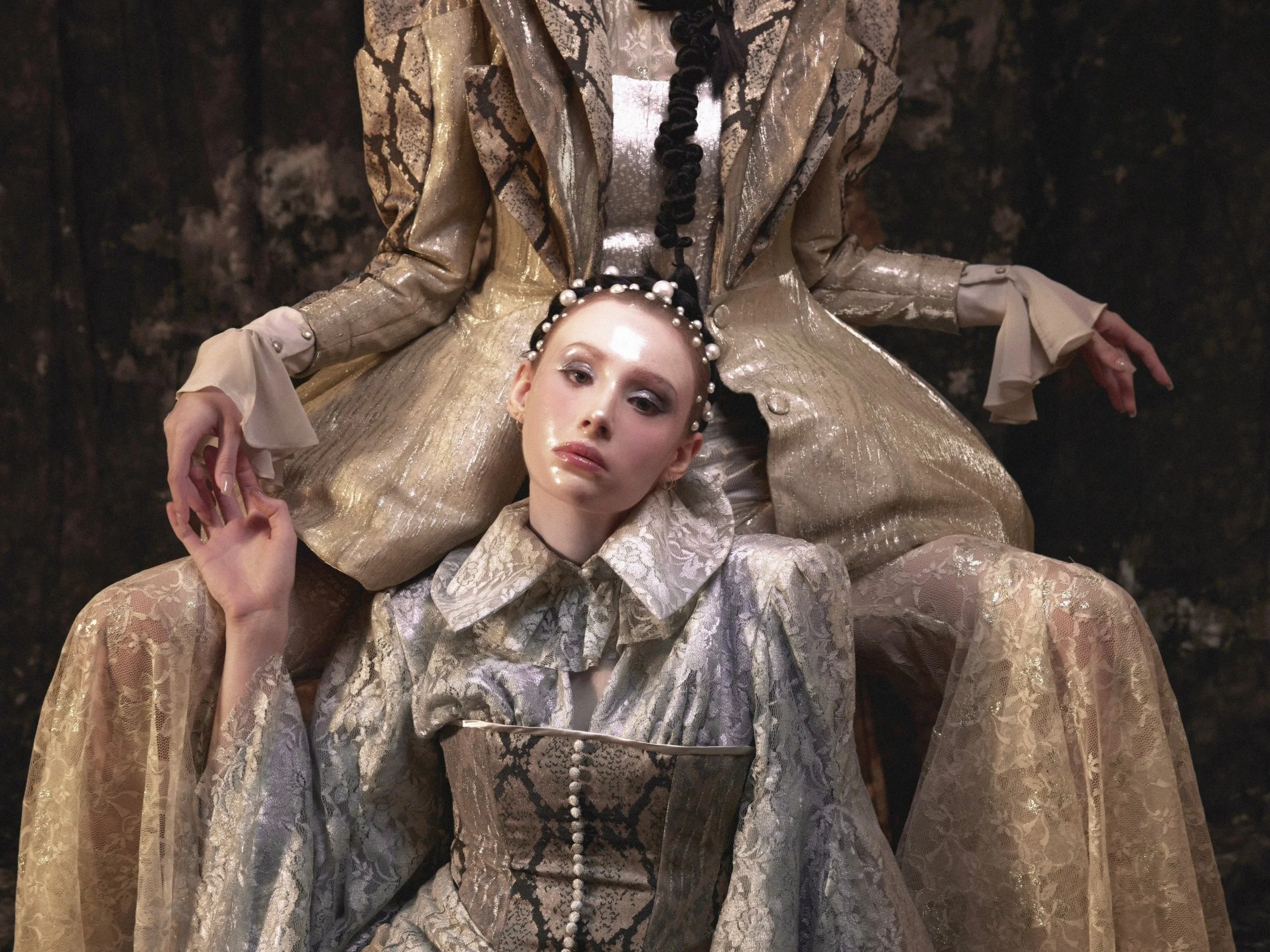 Fashion model in an ornate gold and lace dress with pearl and snake skin pattern details, wearing a pearl headband, standing against a dark background.