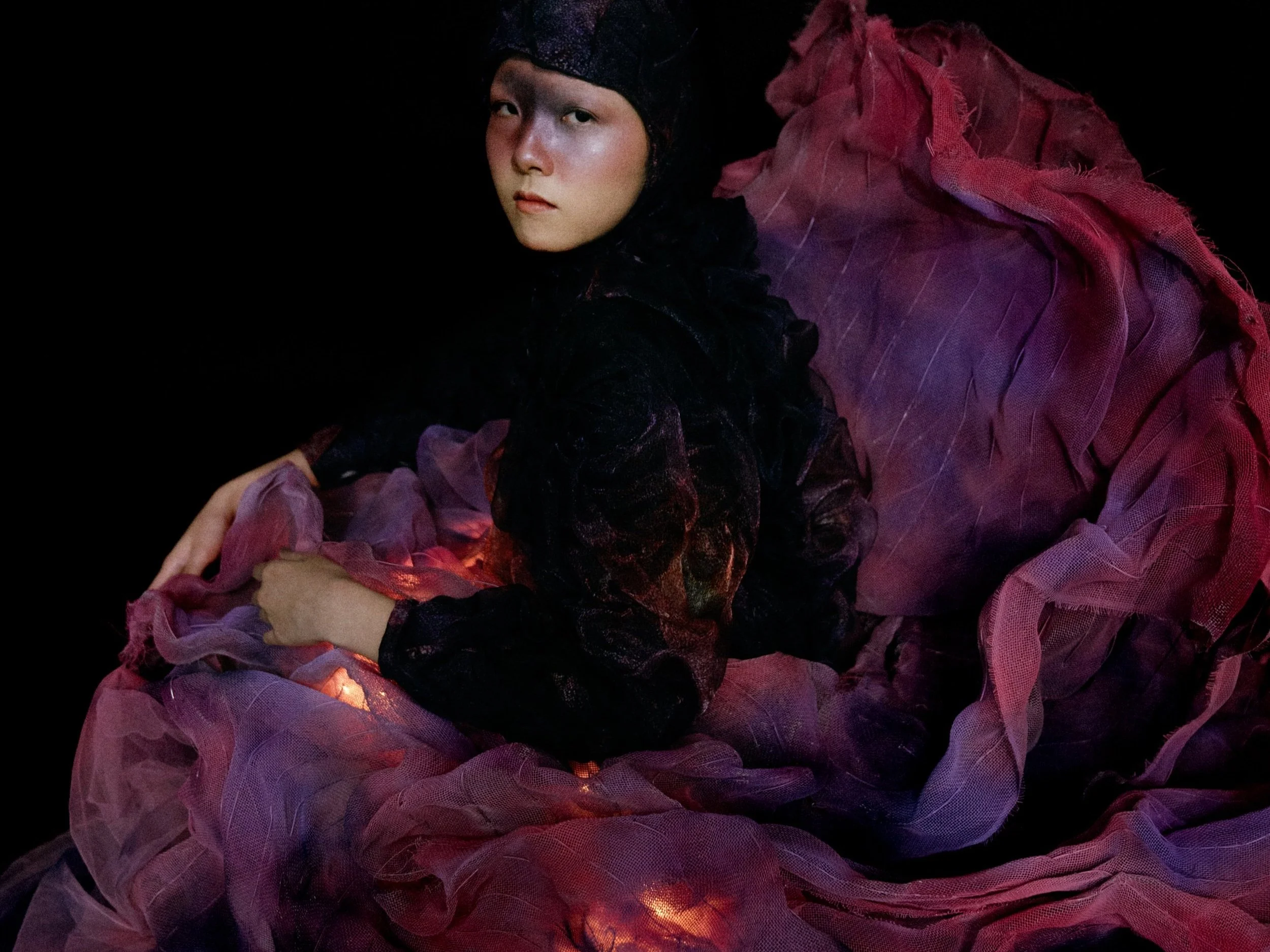 A woman with a neutral expression wearing a black head covering and dark clothing, surrounded by pink and purple mesh fabric, in a dark setting.