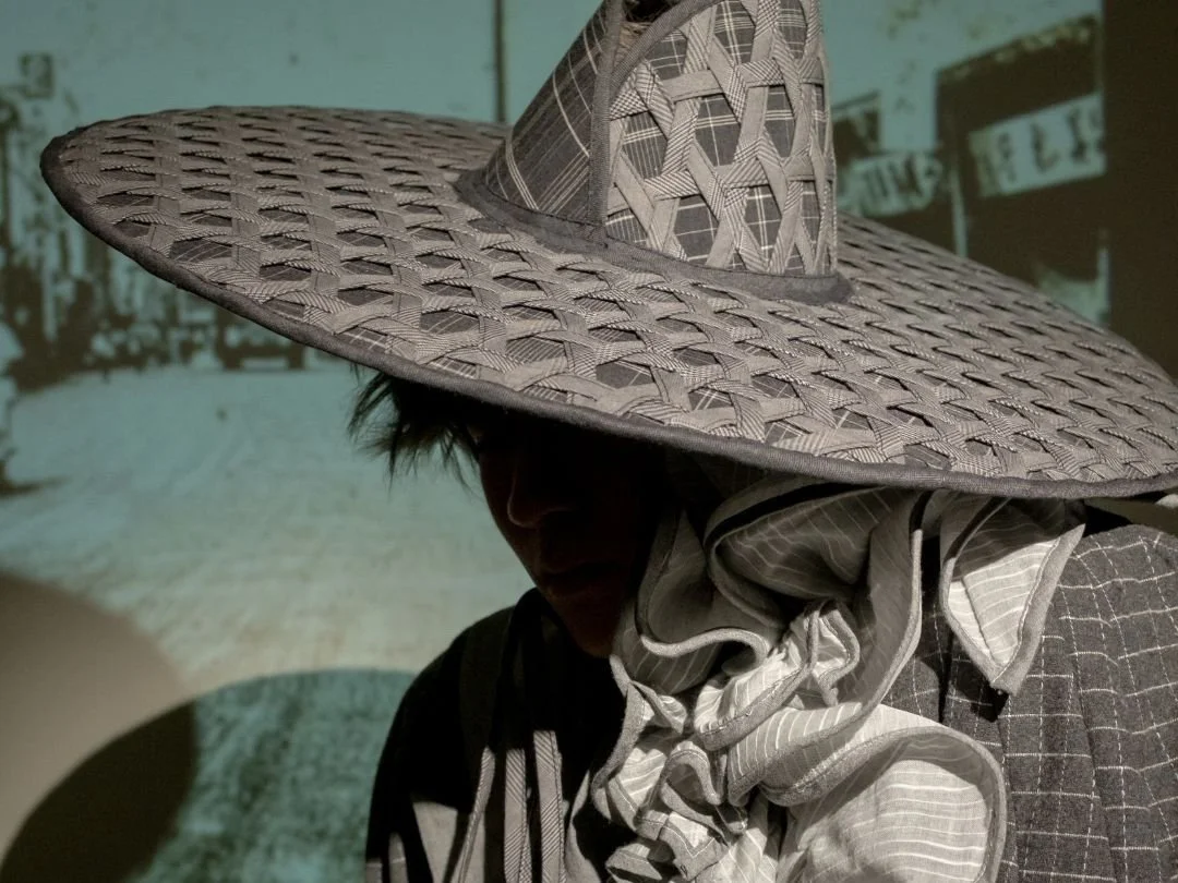 A person wearing a large woven straw hat with a wide brim, a patterned scarf, and clothing with striped and checked patterns, with a blurred urban street scene in the background.