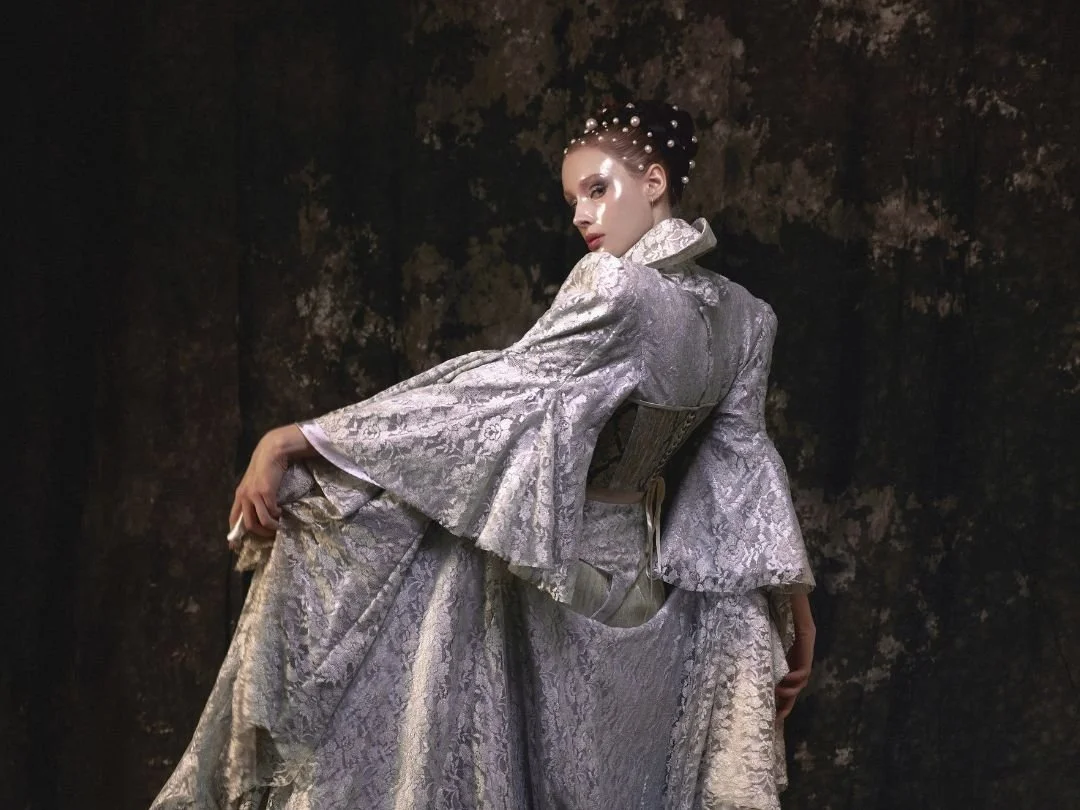 A woman in an elaborate silver-gray brocade gown with flared sleeves, styled with a corset, stands against a dark textured background, with her hair styled in an updo adorned with pearls.