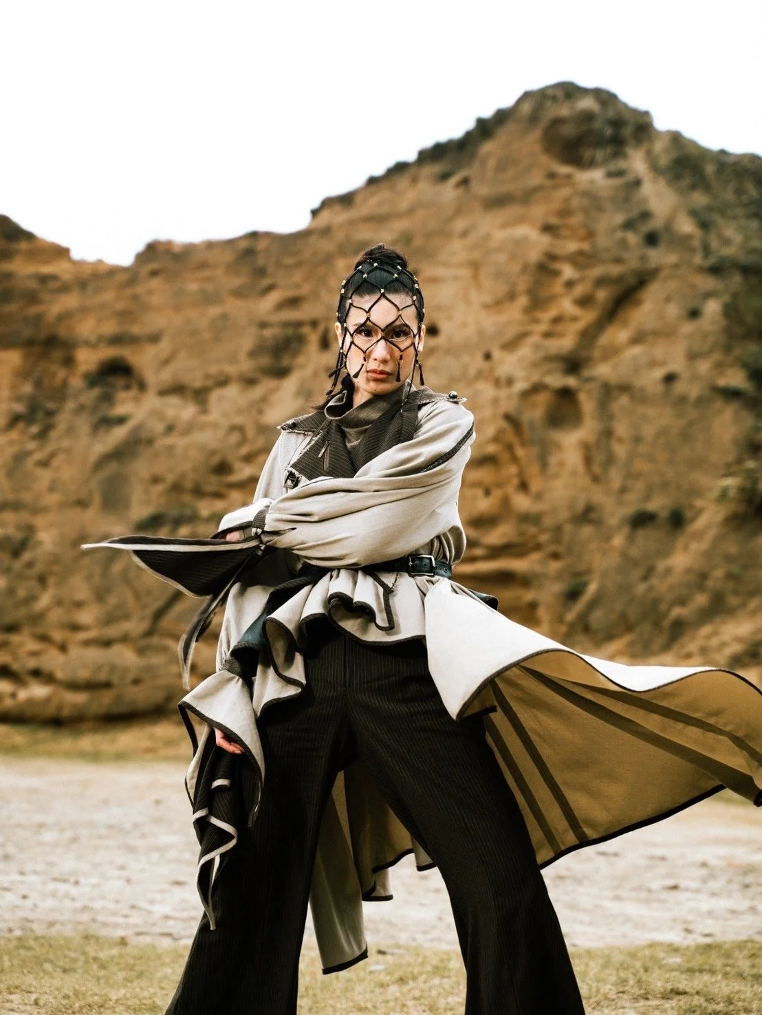Person in avant-garde fashion outfit with face netting, standing outdoors against a rocky landscape.