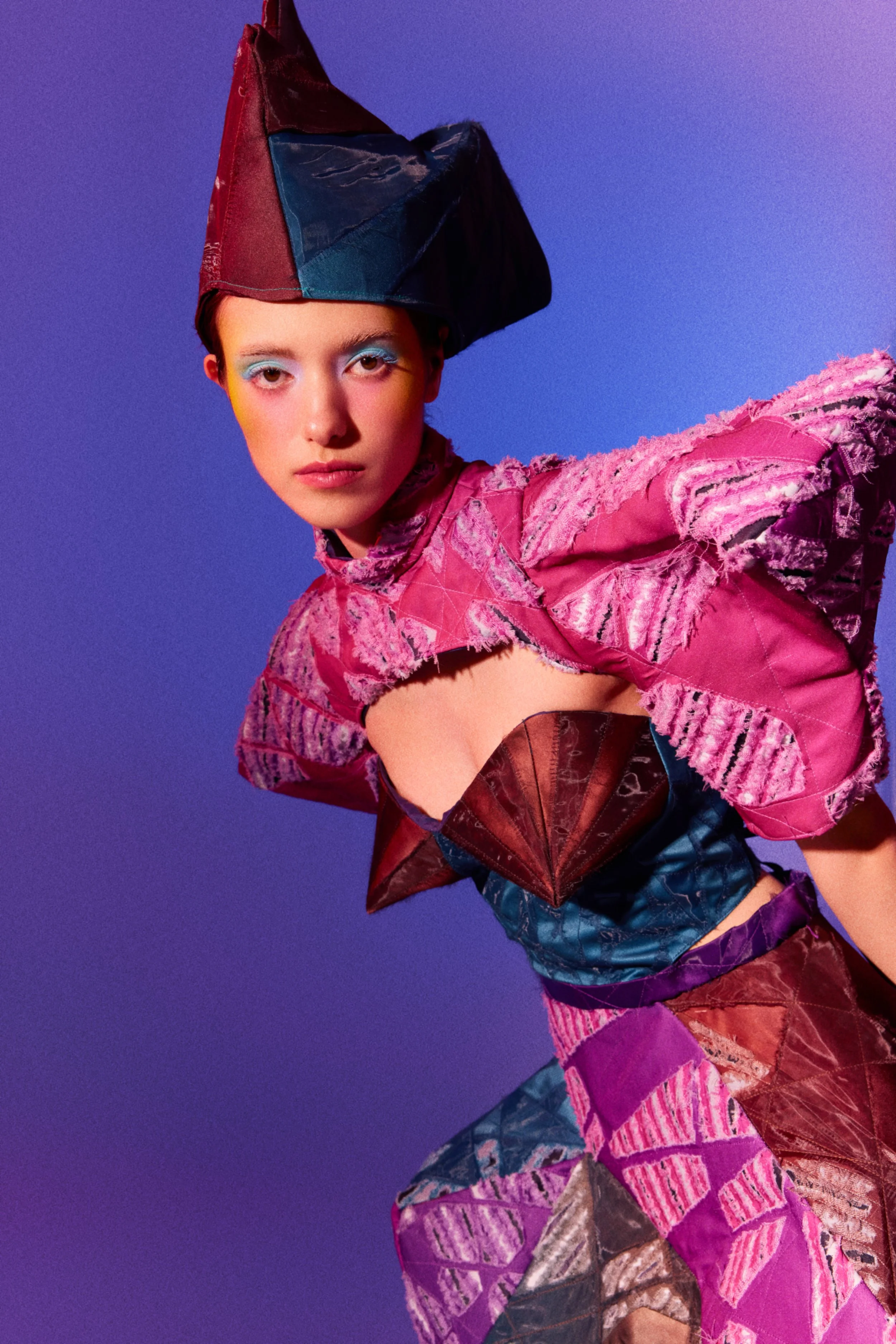 A person with short hair wearing vibrant, colorful clothing made of patchwork fabrics in pink, purple, red, and brown tones. They have bold makeup with blue eyeshadow and are posed against a purple gradient background, with their arm extended forward.