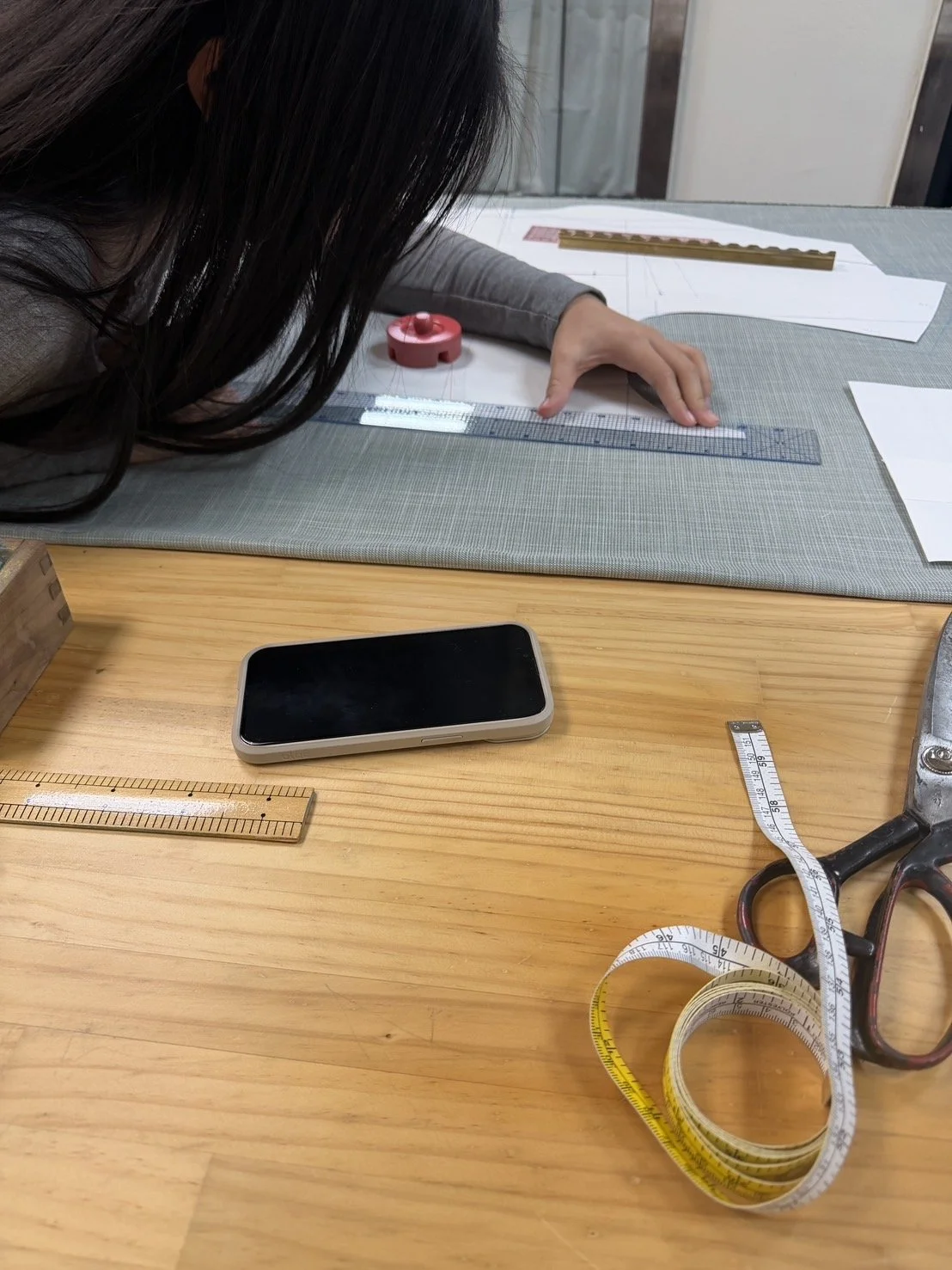 A person working on a sewing or design project at a table with various measuring tools, a smartphone, and sewing patterns.