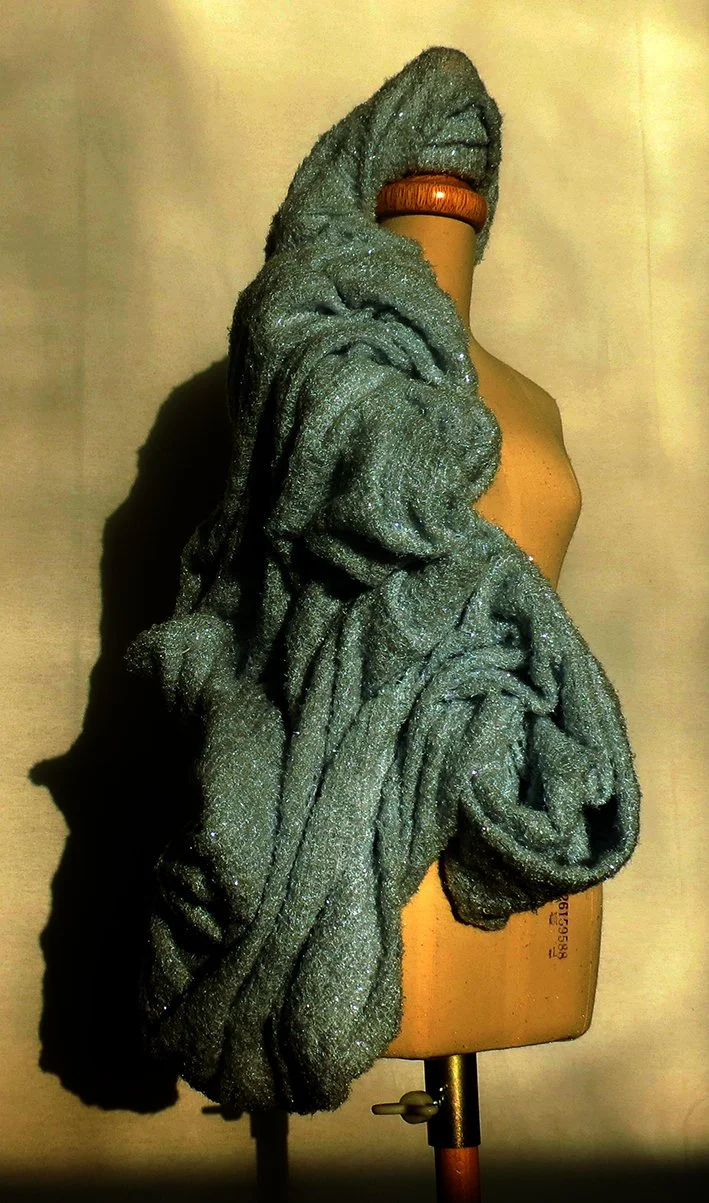 A mannequin with a beige torso and wooden neck, draped with a sparkly gray knitted scarf that extends over the shoulder.