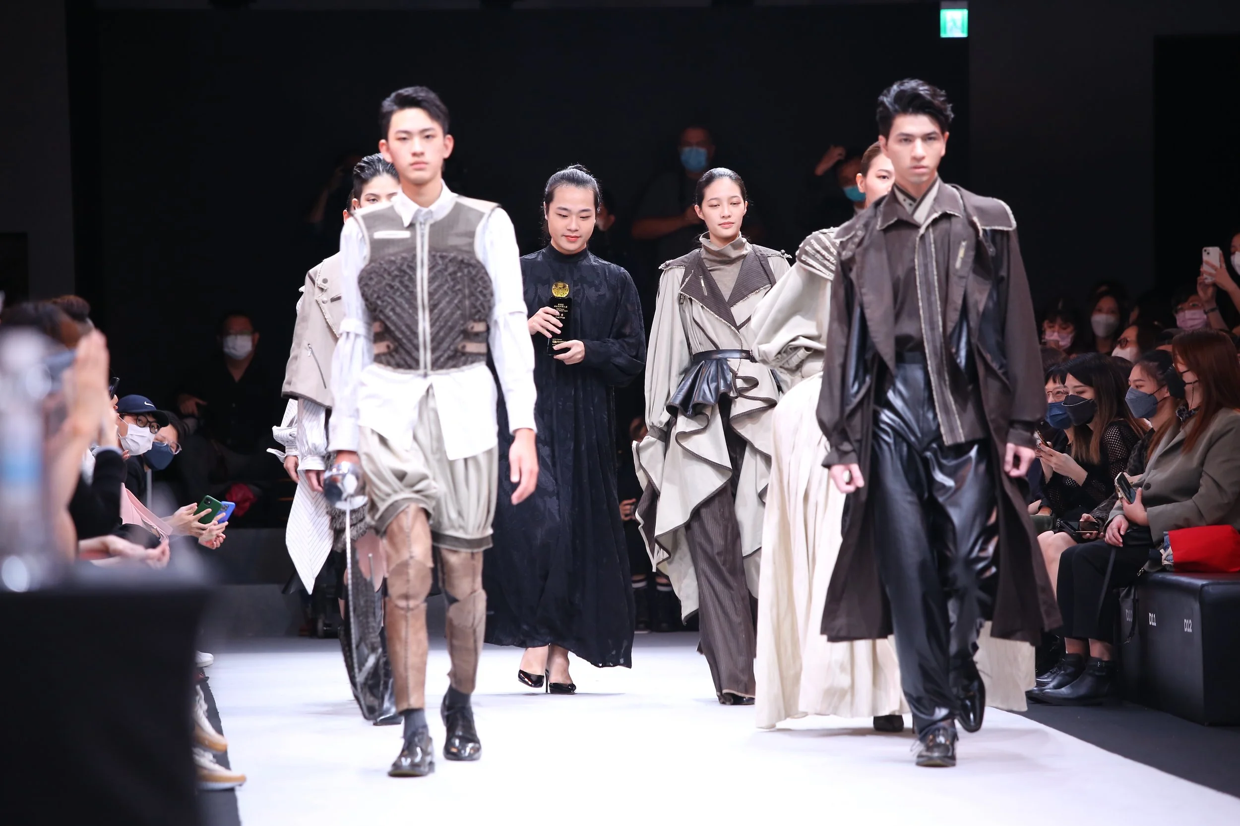 Models walking down a runway during a fashion show with an audience seated on both sides, some wearing masks and taking photos.