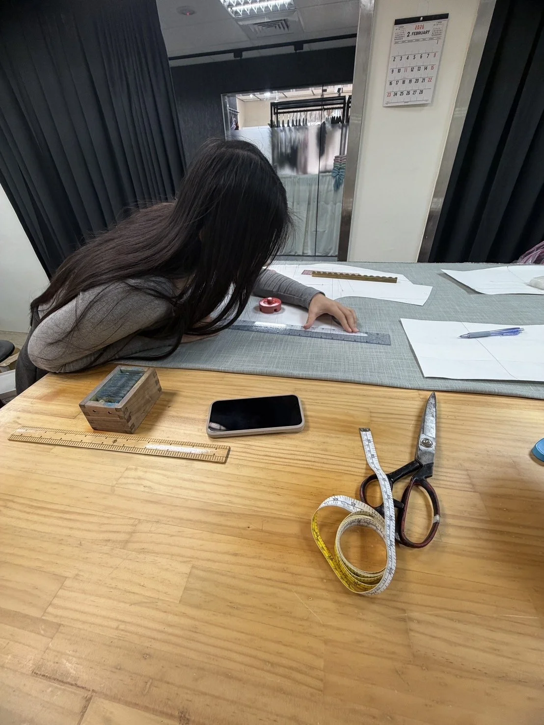 A woman measuring a fabric using a ruler on a cutting mat. On the table, there are scissors, a smartphone, a carpenter's square, a wooden box, pens, paper patterns, and a tape measure. In the background, a calendar is hanging on the wall, and a mirro