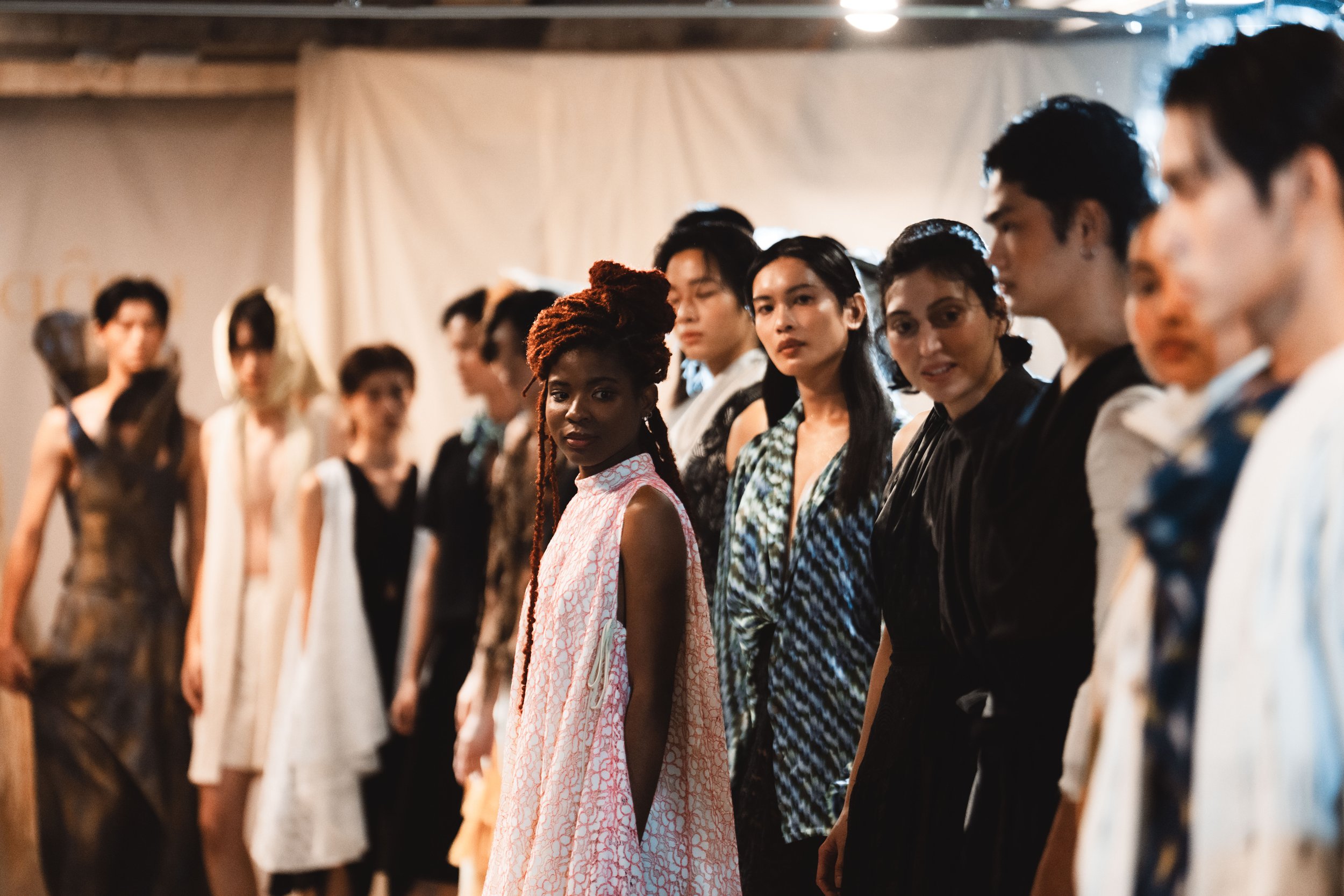 A group of models on a runway, wearing various stylish outfits. The focus is on a woman with braided hair wearing a pink dress, with other models standing alongside her. The background is blurred.
