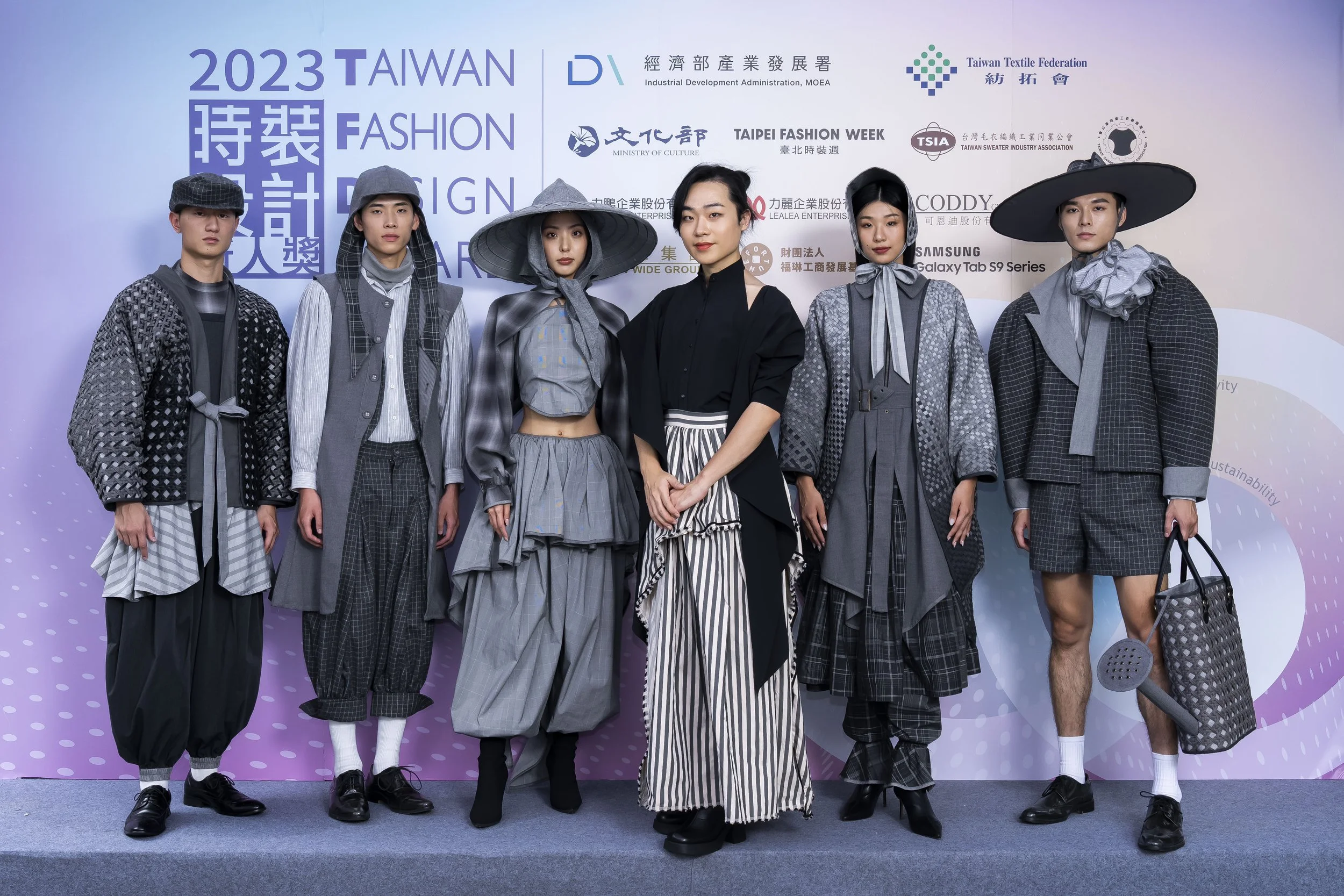 Group of six models wearing trendy, eclectic clothing and accessories, posing in front of a purple backdrop with event and sponsor logos including Taiwan Textile Federation and Taipei Fashion Week, at the 2023 Taiwan Fashion Design Awards.