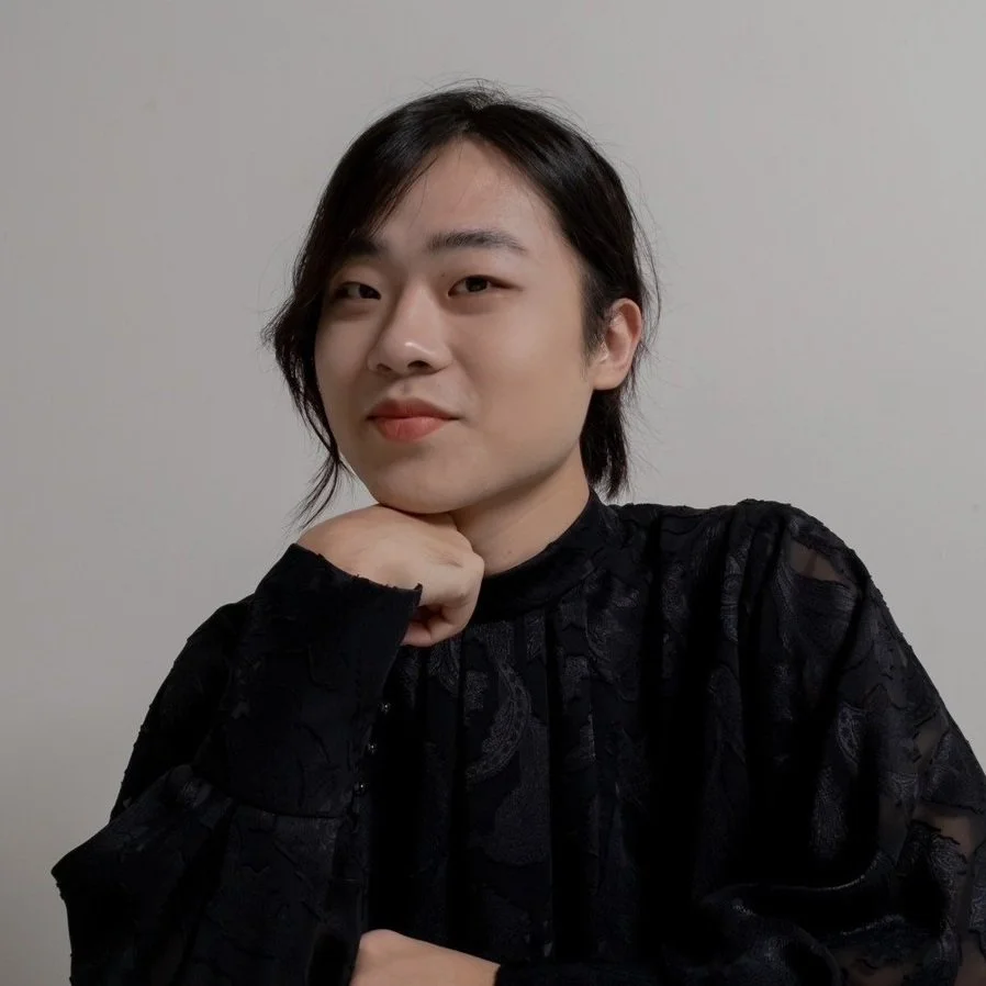 A person with short black hair and light skin, wearing a black textured top, resting their chin on their hand, and looking at the camera with a slight smile, against a plain gray wall.
