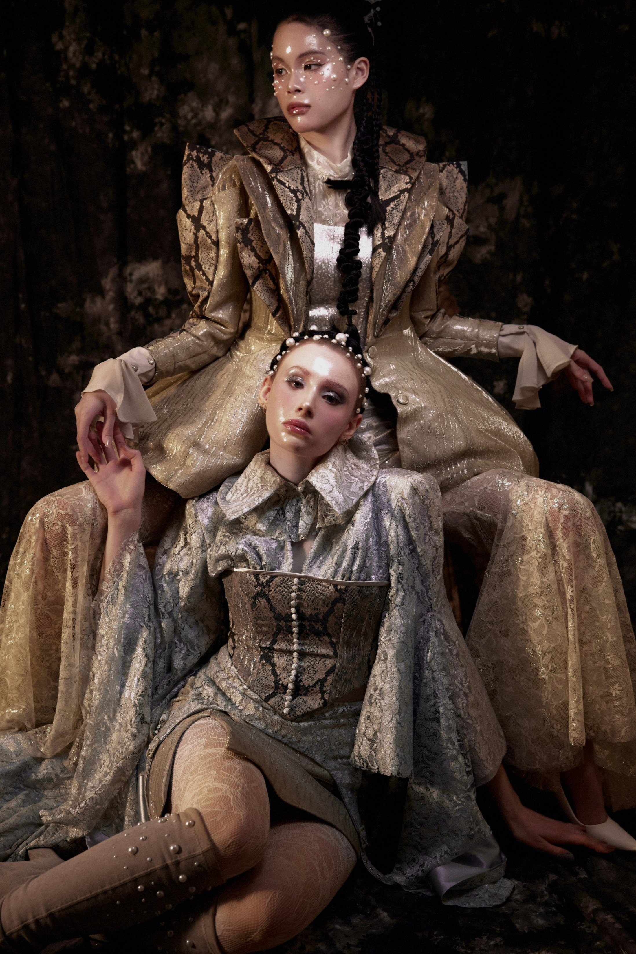 Two women dressed in elaborate, gold and snake-print outfits with pearls and lace, with one woman sitting on the other's lap, posing against a dark background.