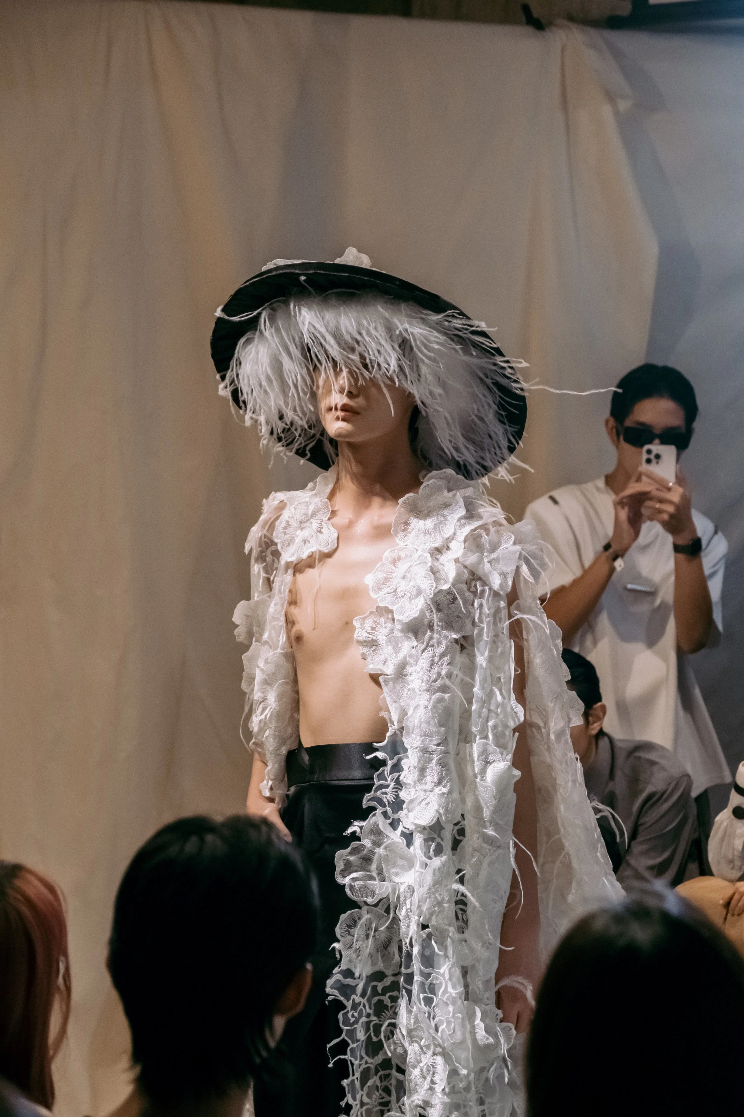 A fashion model wearing an intricate, sheer outfit with floral details and a large feathered hat on a runway, while an audience watches and photographs the scene.