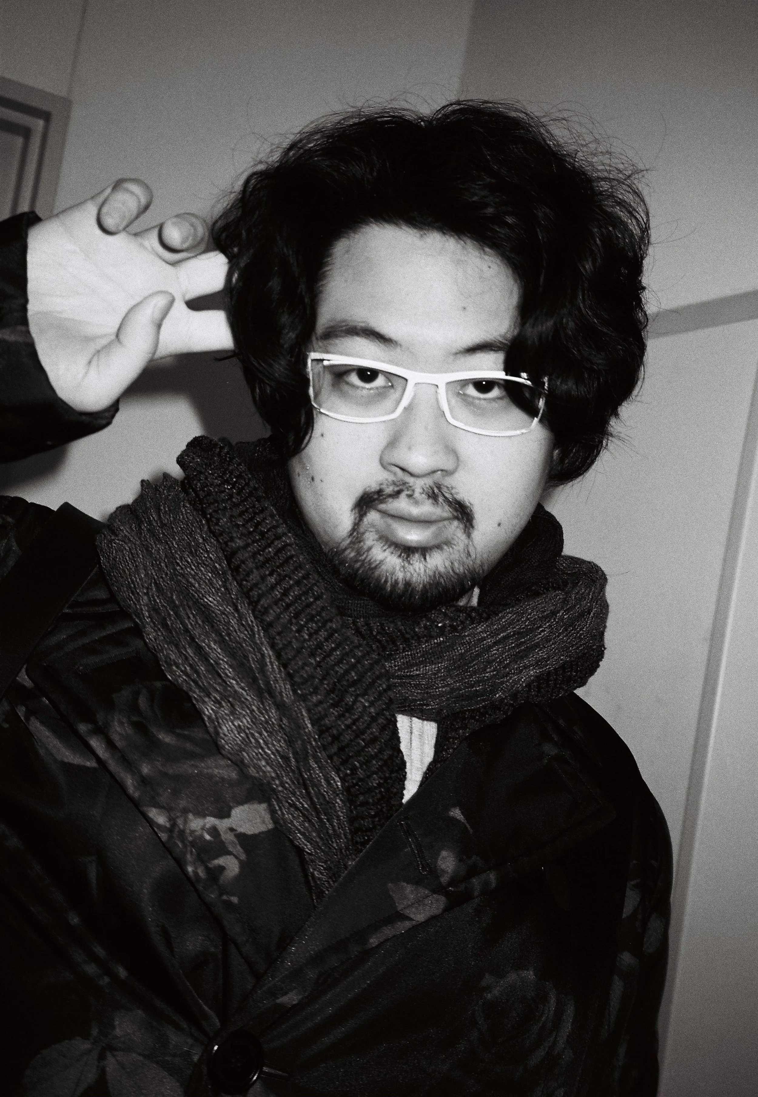 A person with curly dark hair wearing glasses and a dark jacket, with a scarf, raising their hand near their head inside a room with plain walls.