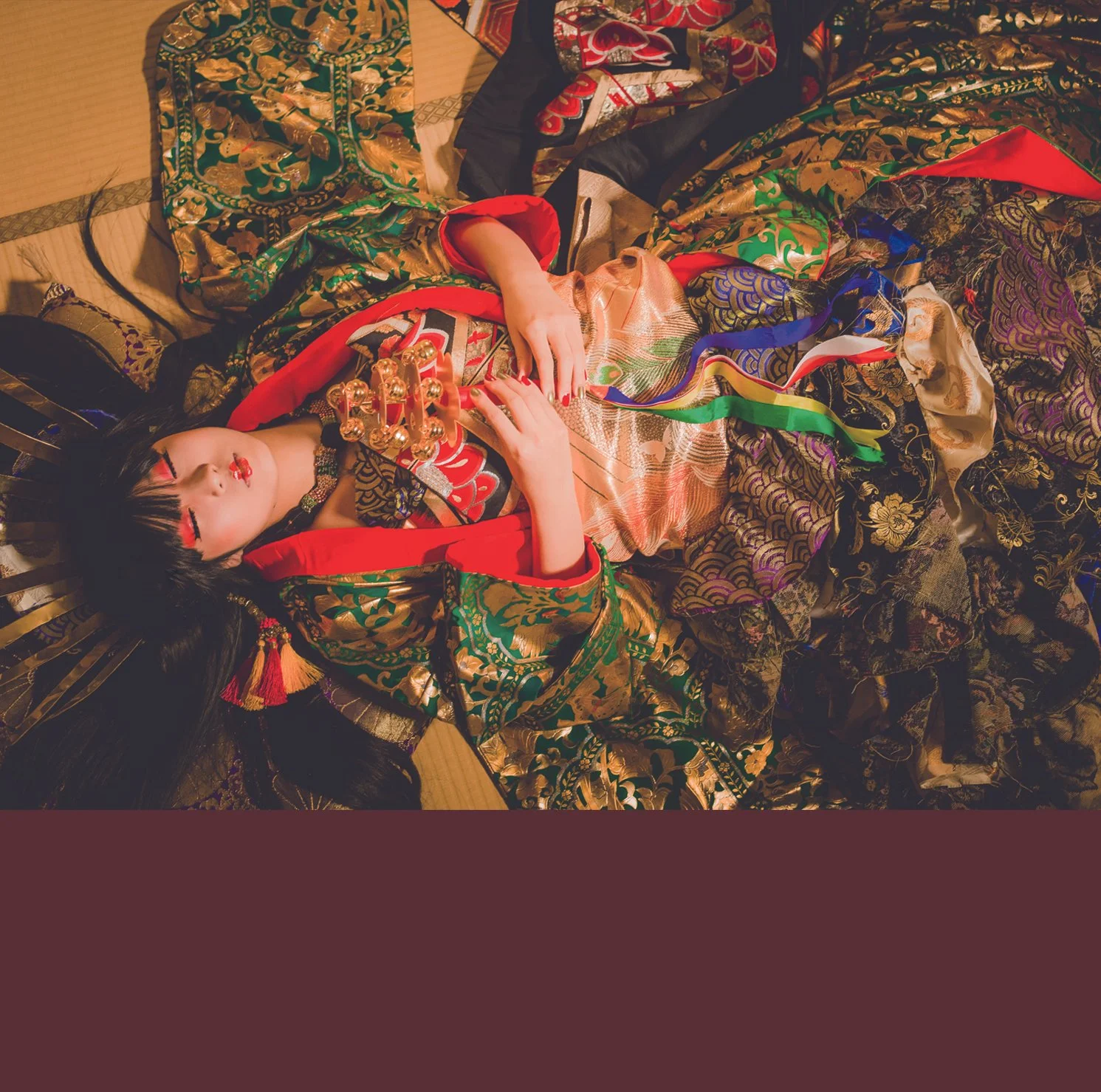 A woman dressed in a colorful, ornate kimono lying on the floor with her eyes closed, surrounded by decorative fabrics and traditional Japanese items.