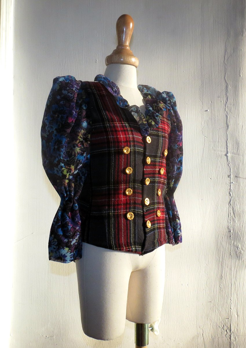 A mannequin wearing a vintage-style blazer with a plaid pattern in red, black, and yellow, adorned with gold buttons, and featuring floral-patterned puffy sleeves in blue and purple.