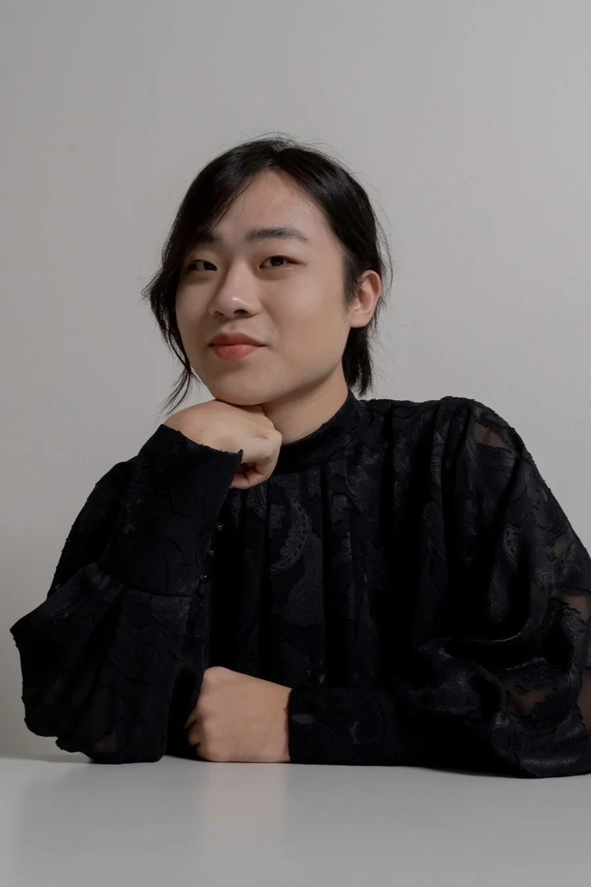 Portrait of a young person with short dark hair, wearing a black patterned lace top, sitting at a white table, resting their chin on their hand, against a plain light-colored wall.