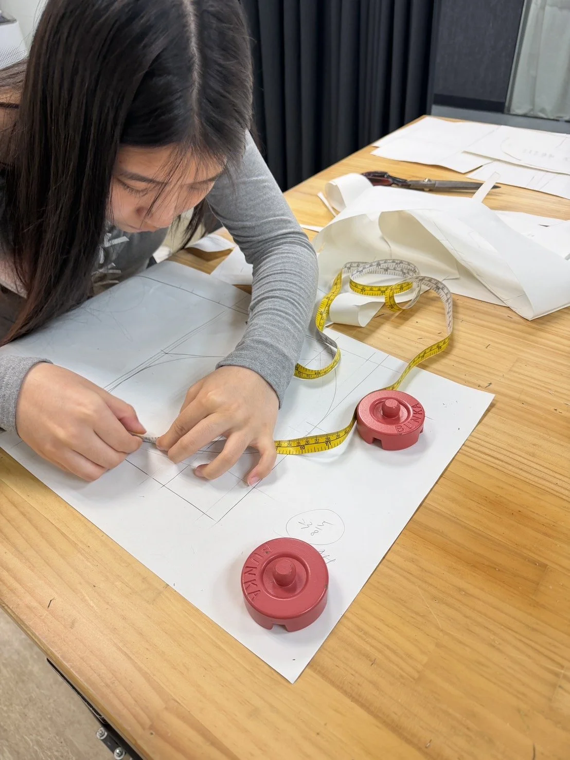 A woman is working on a detailed fashion sketch or pattern design on large white paper. She is using a measuring tape and a pencil, with additional papers and pens spread out on the wooden table.