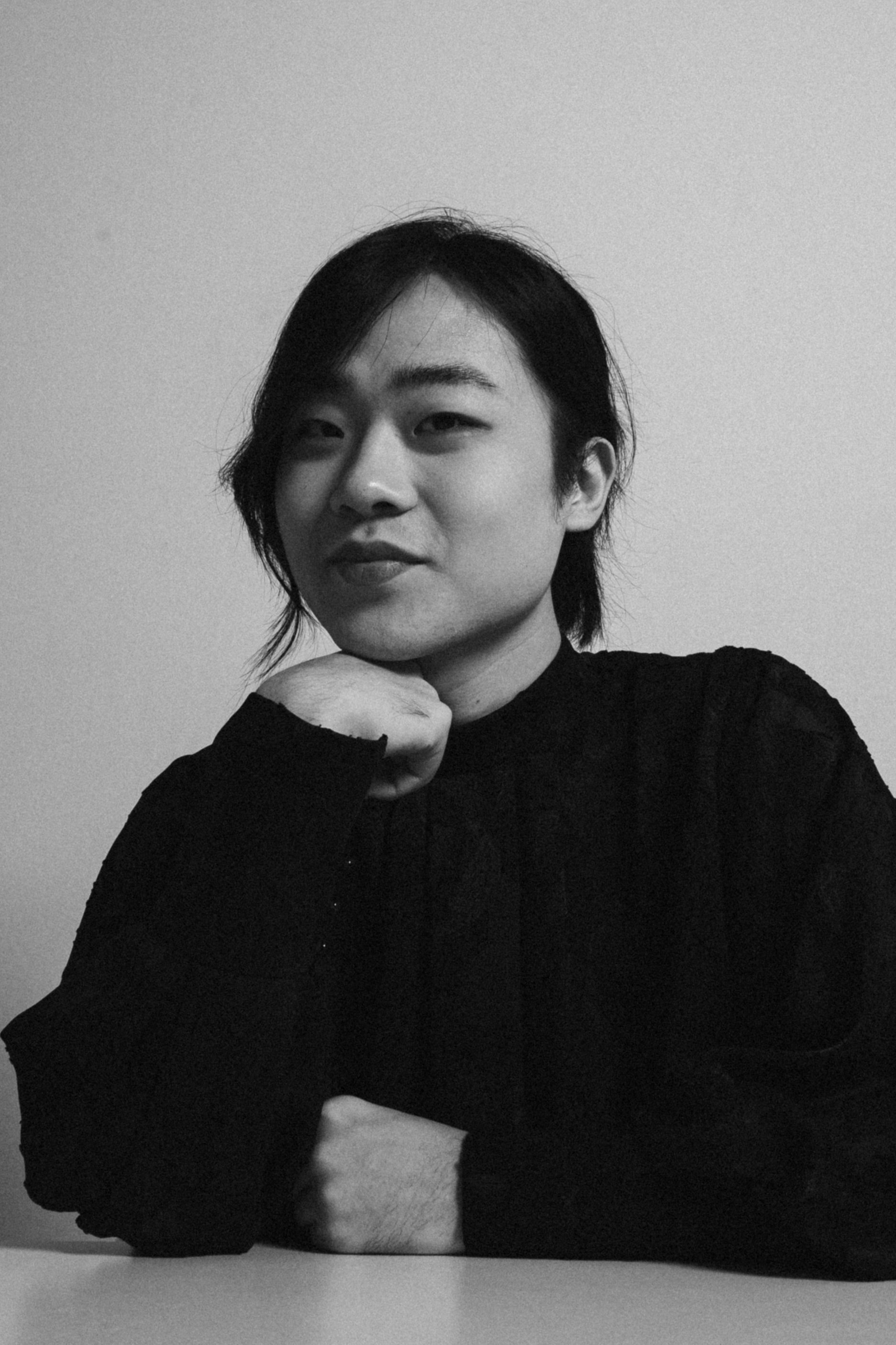 Black and white photo of a woman with short hair, resting her chin on her hand, sitting at a table, with a neutral background.