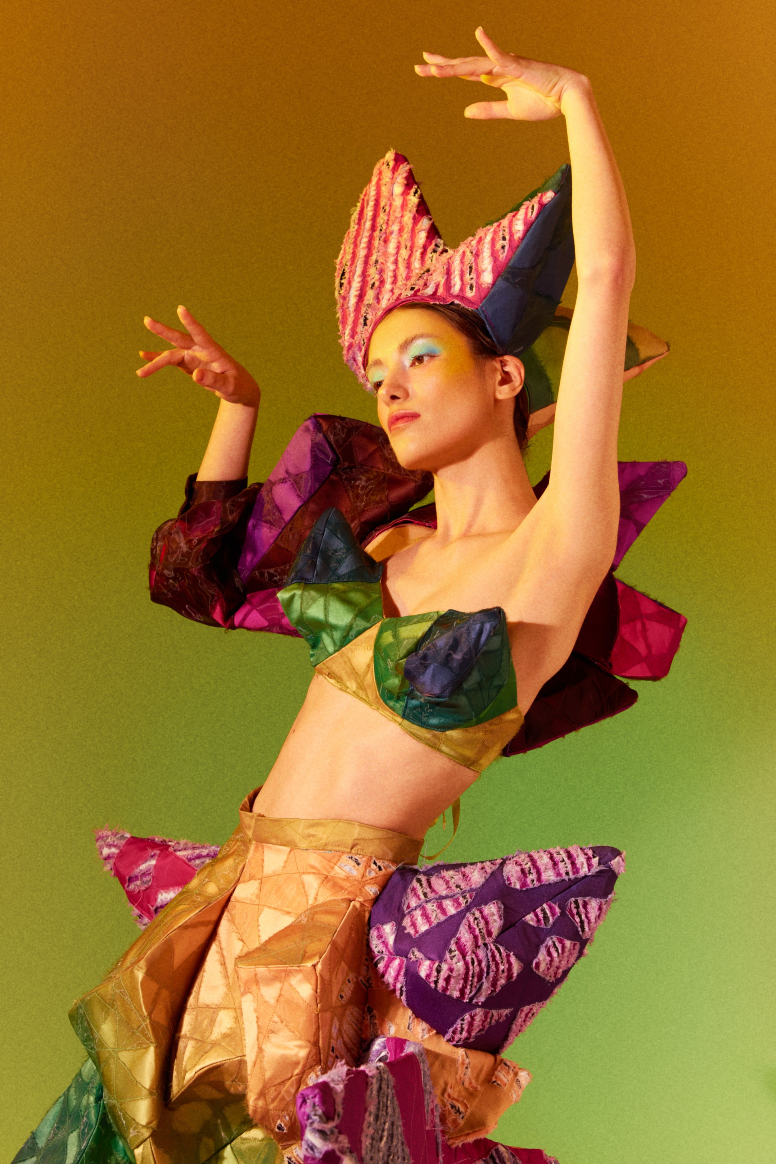 A woman wearing a colorful, avant-garde costume with textured, layered fabric and a tall, elaborate headpiece, striking a pose against a plain green background.