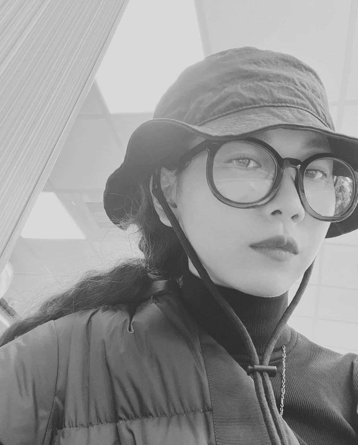 Black and white photo of a young woman wearing glasses, a wide-brimmed hat, and a puffer jacket, looking at the camera.