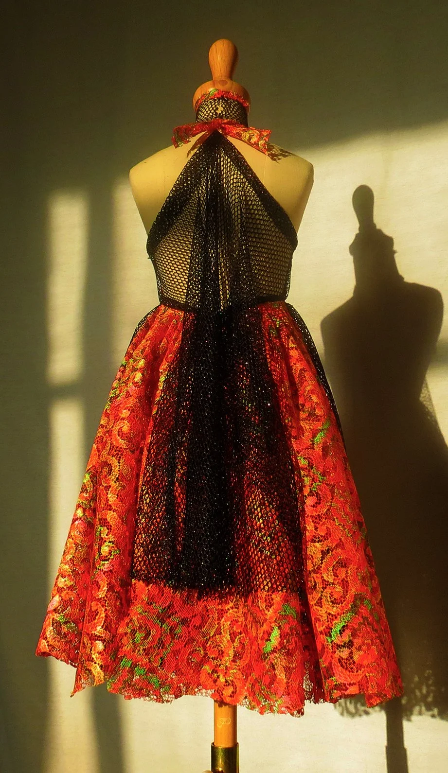 Fashion dress on a mannequin with black lace overlay, red and sparkly fabric, and a red ribbon tied around the neck. Shadow cast on wall.