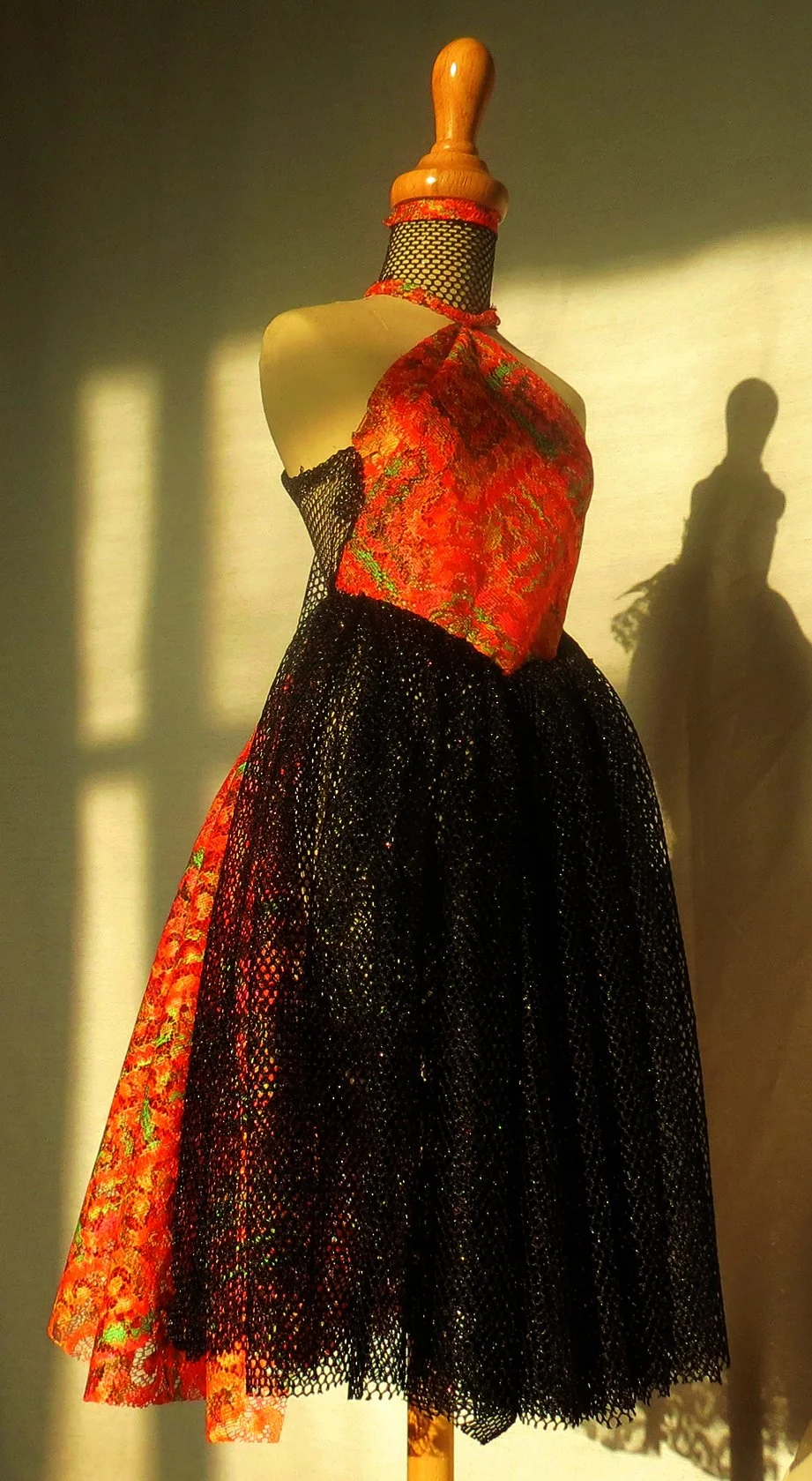 A mannequin displays a vintage dress with a red and gold floral bodice and a black mesh princess-style skirt, illuminated by sunlight casting shadows.