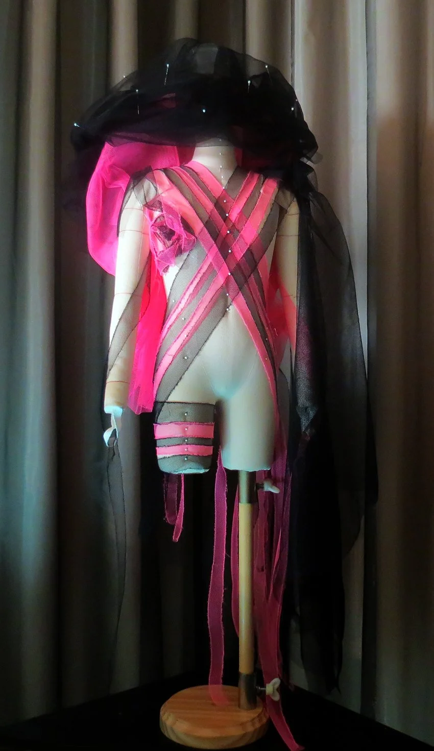A mannequin dressed in pink and black lingerie with sheer fabric and black veil. The display includes various straps and pink ribbons, with a black veil draped over the head.