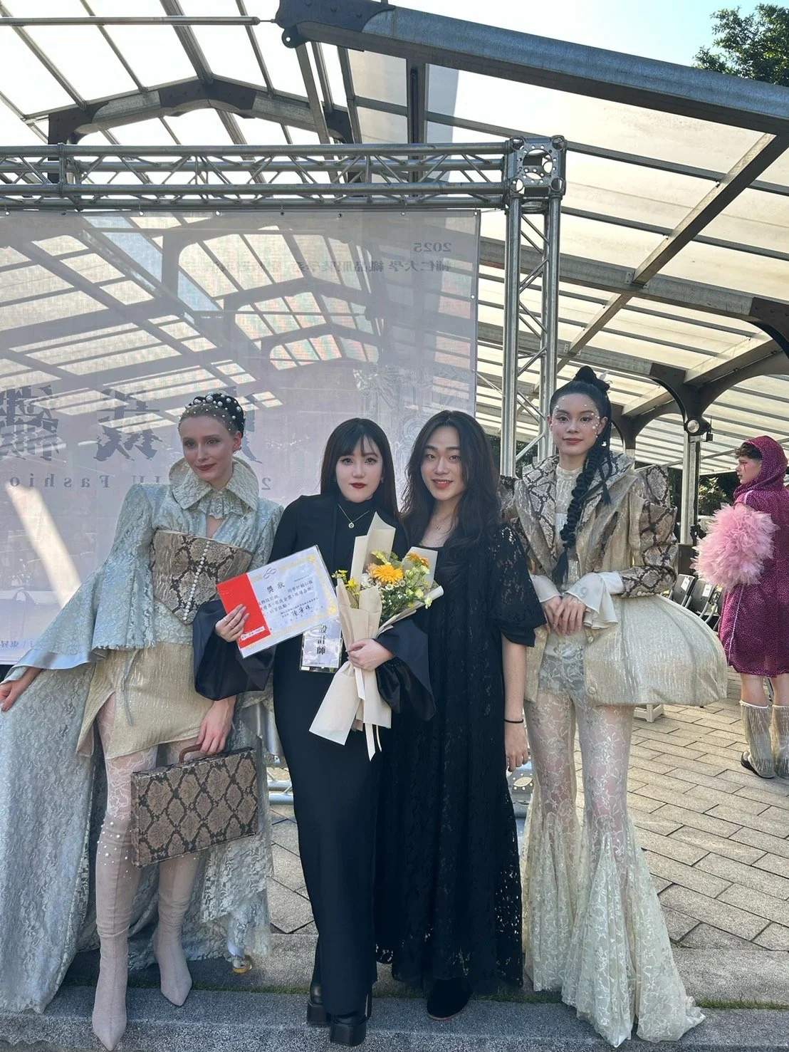 Four women, one holding a bouquet of flowers, posing together at a graduation or award event outdoors under a glass roof.
