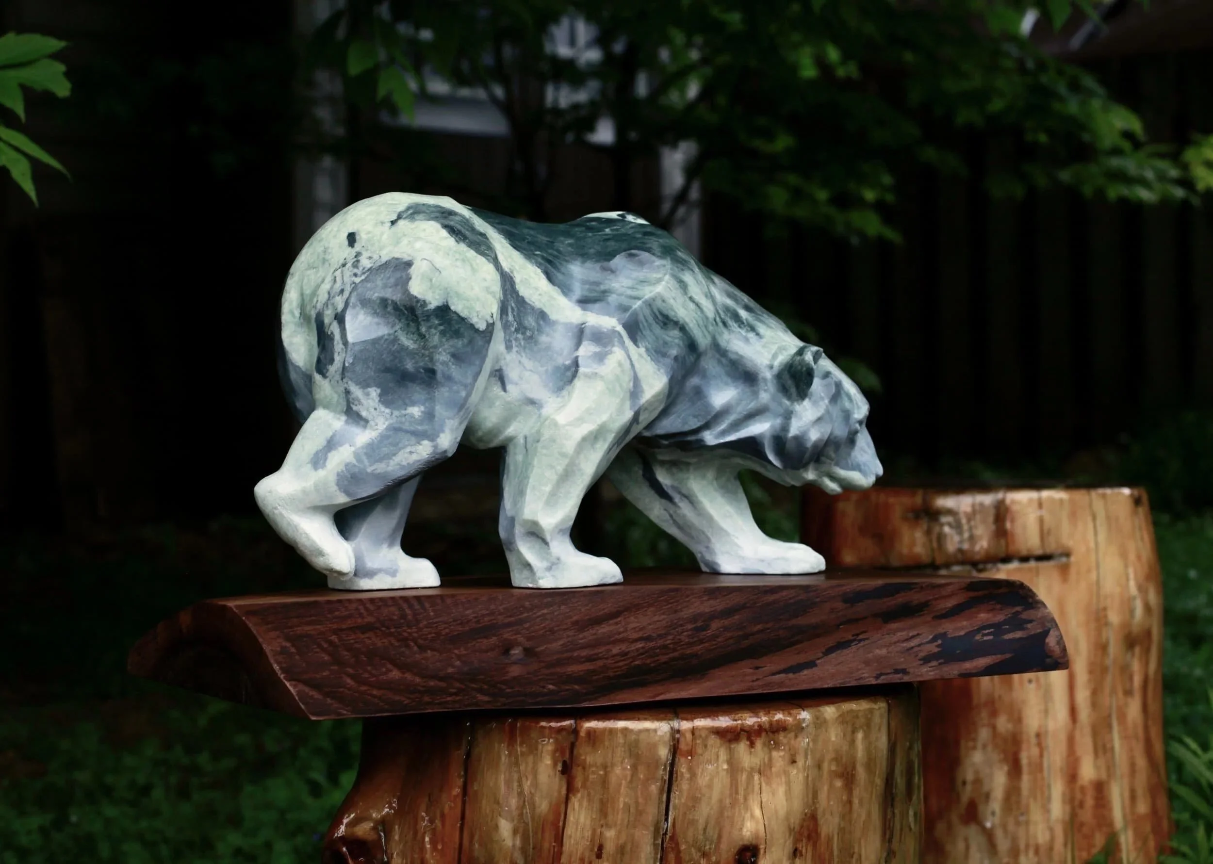 A carved stone bear sculpture stands on a wooden slab, displayed outdoors on top of a tree stump.