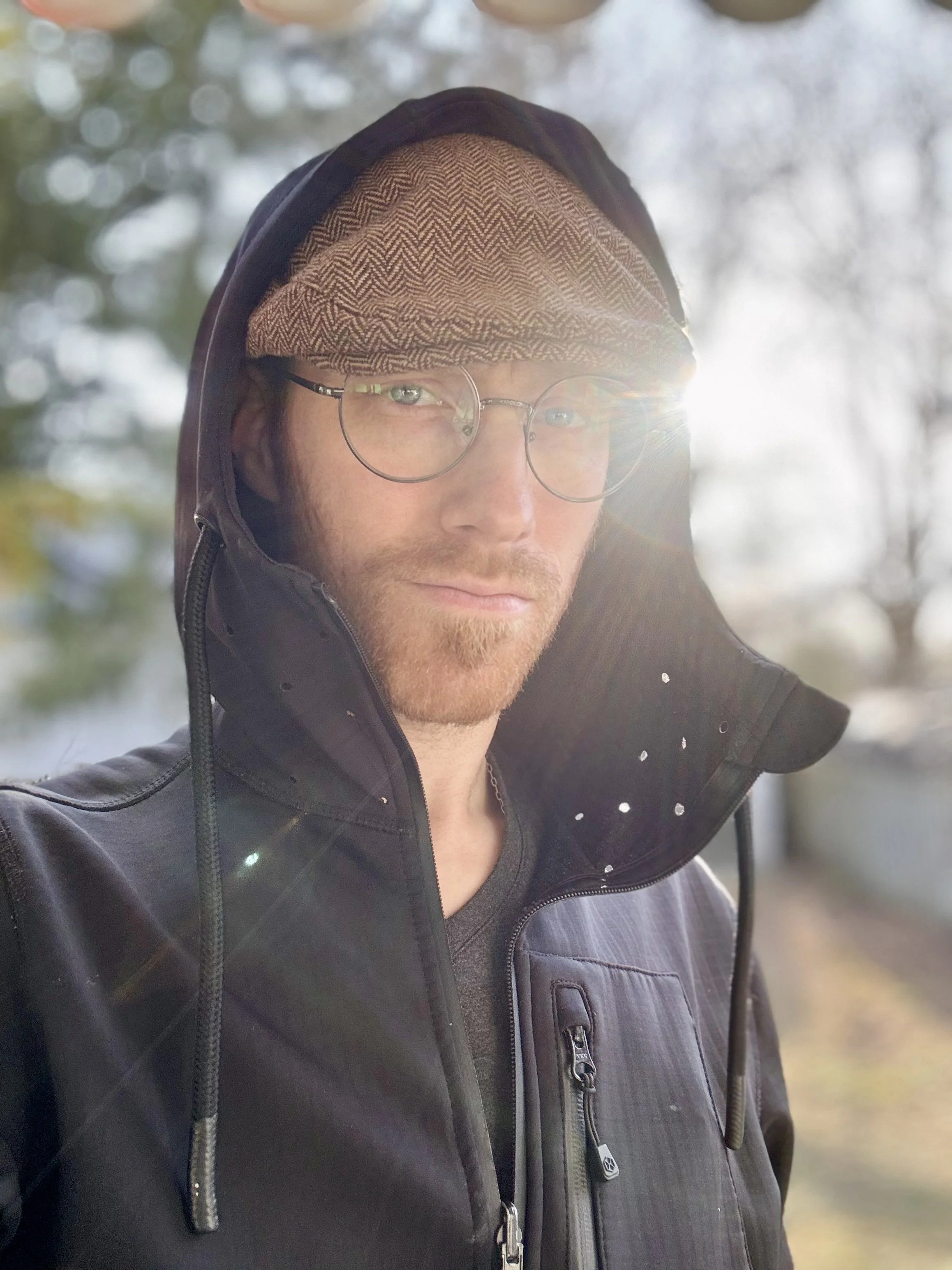 Person wearing glasses and a brown flat cap, with a black hoodie, standing outdoors in sunlight - Wesley Booker