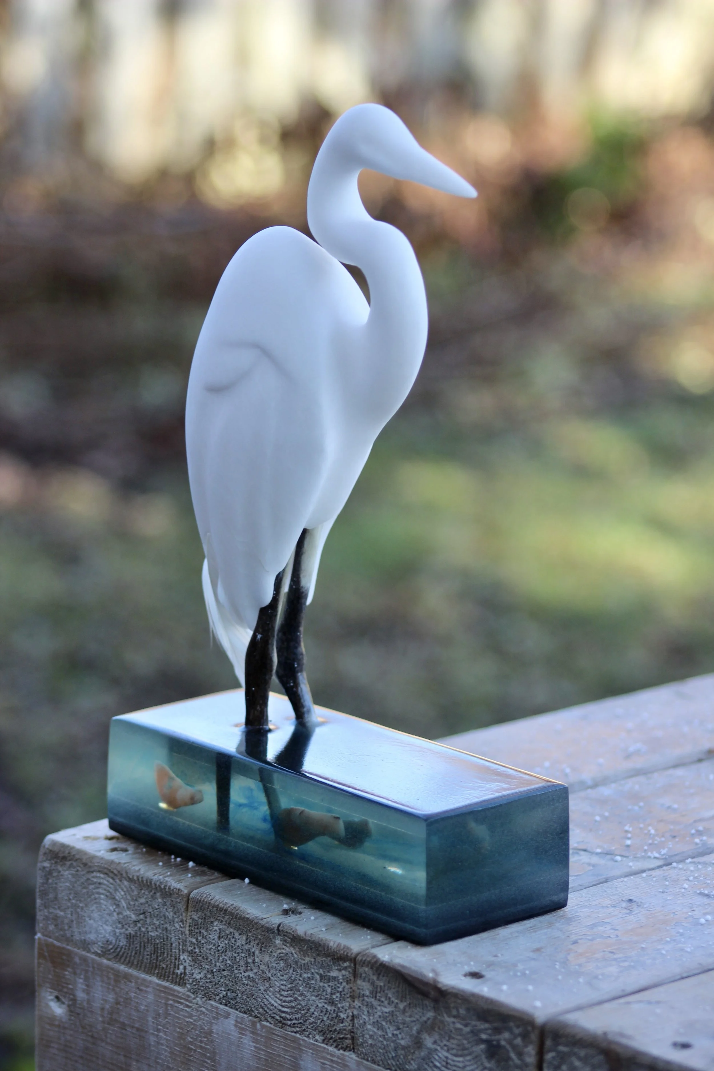 Decorative alabaster sculpture of a white heron with elk horn legs standing on an epoxy base with alabaster fish inside, placed on a wooden surface outdoors.  Made by Wesley Booker 2023.