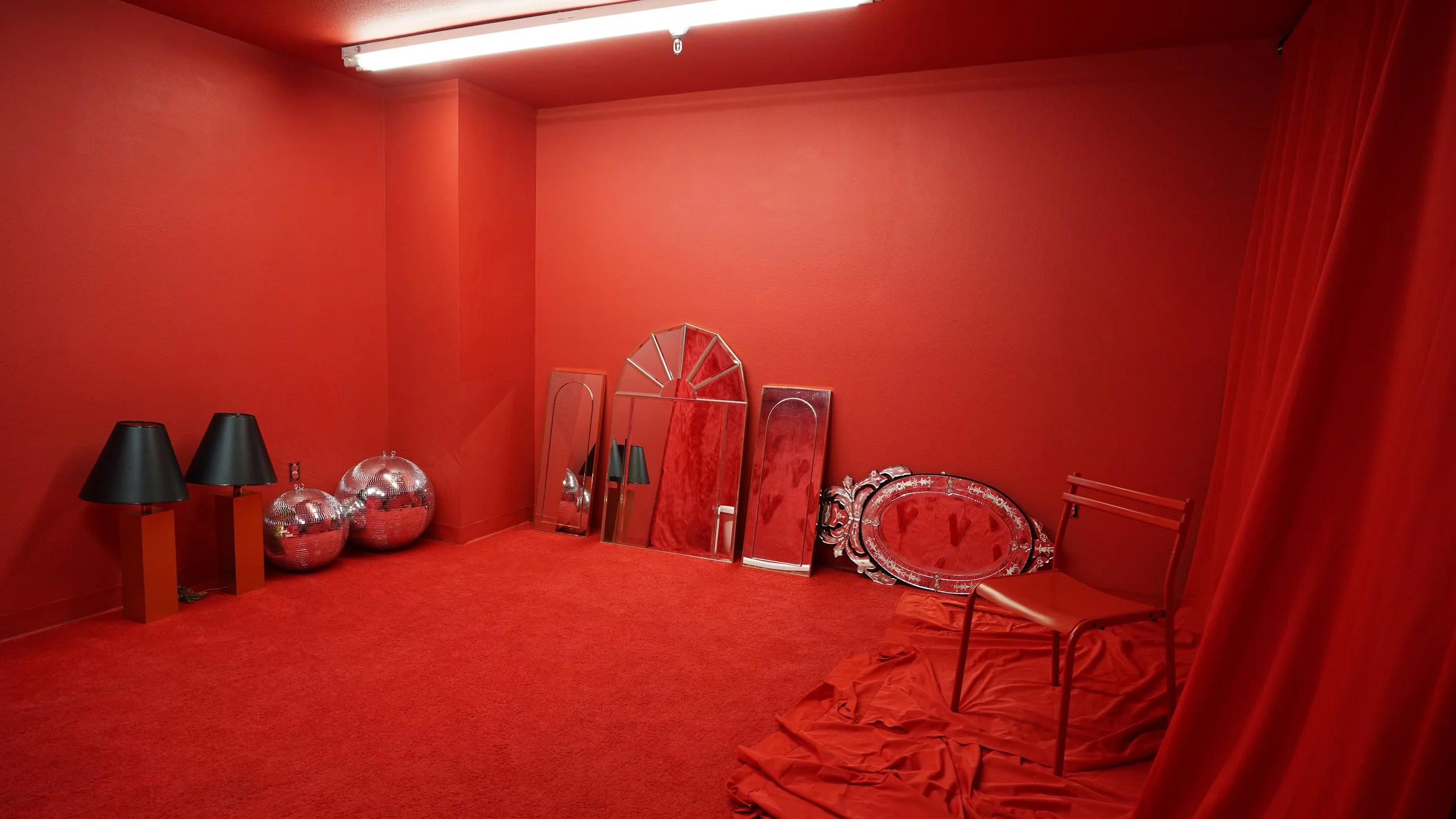 A room with red walls and a red carpet, featuring two black lamps with black shades on orange stands, two silver disco balls, a collection of three tall mirrors leaning against the wall, one of which is arched, a decorative ornate silver frame, a red