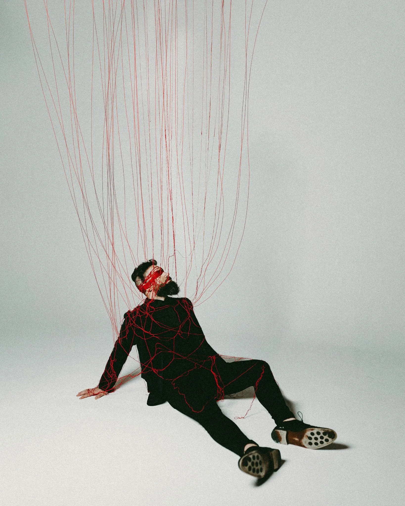 A woman with dark hair and glasses is dressed in black and sitting on the floor. She is wrapped in red strings that are connected to a large, complex web of red strings above her, with some strings wrapped around her head and body. The background is 