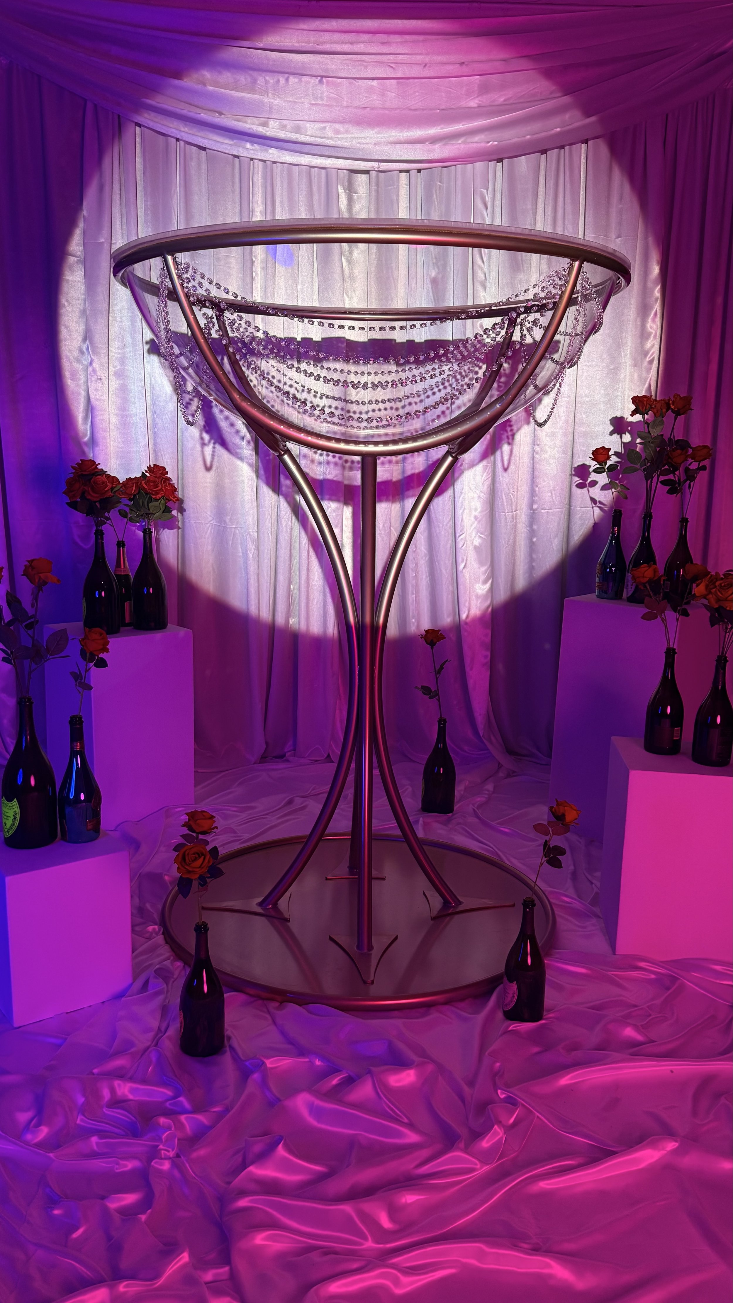 Decorative setting with a large martini glass sculpture decorated with pearls, surrounded by vases of red roses, in a pink and purple lit environment with satin fabric on the floor and backdrop.