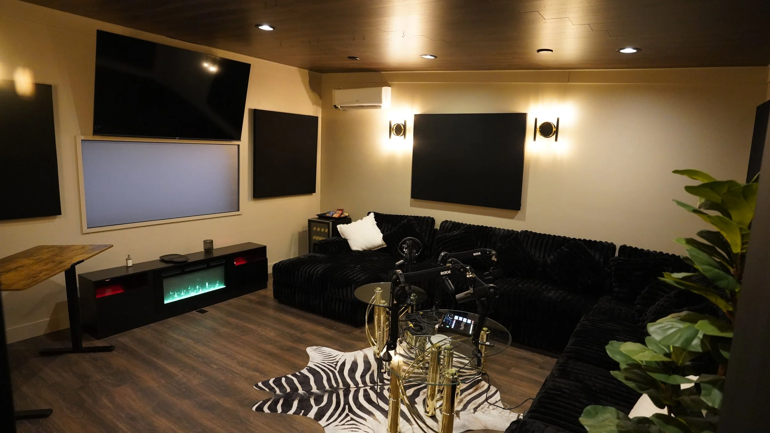 A dimly lit media room with four black wall-mounted screens, a black sectional sofa, a zebra print rug, and a gold-accented coffee table with audio and streaming equipment. A white pillow is on the sofa, and a small side table with snacks is beside i