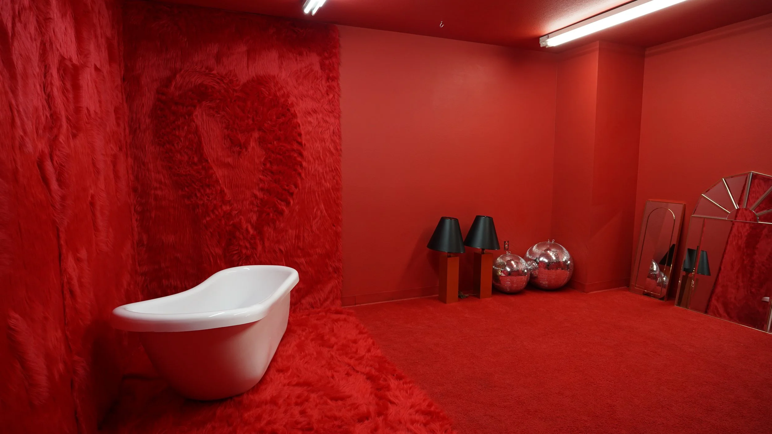 A room with red textured walls and floor, featuring a white bathtub, black lamps on small tables, and silver spheres and mirrors.