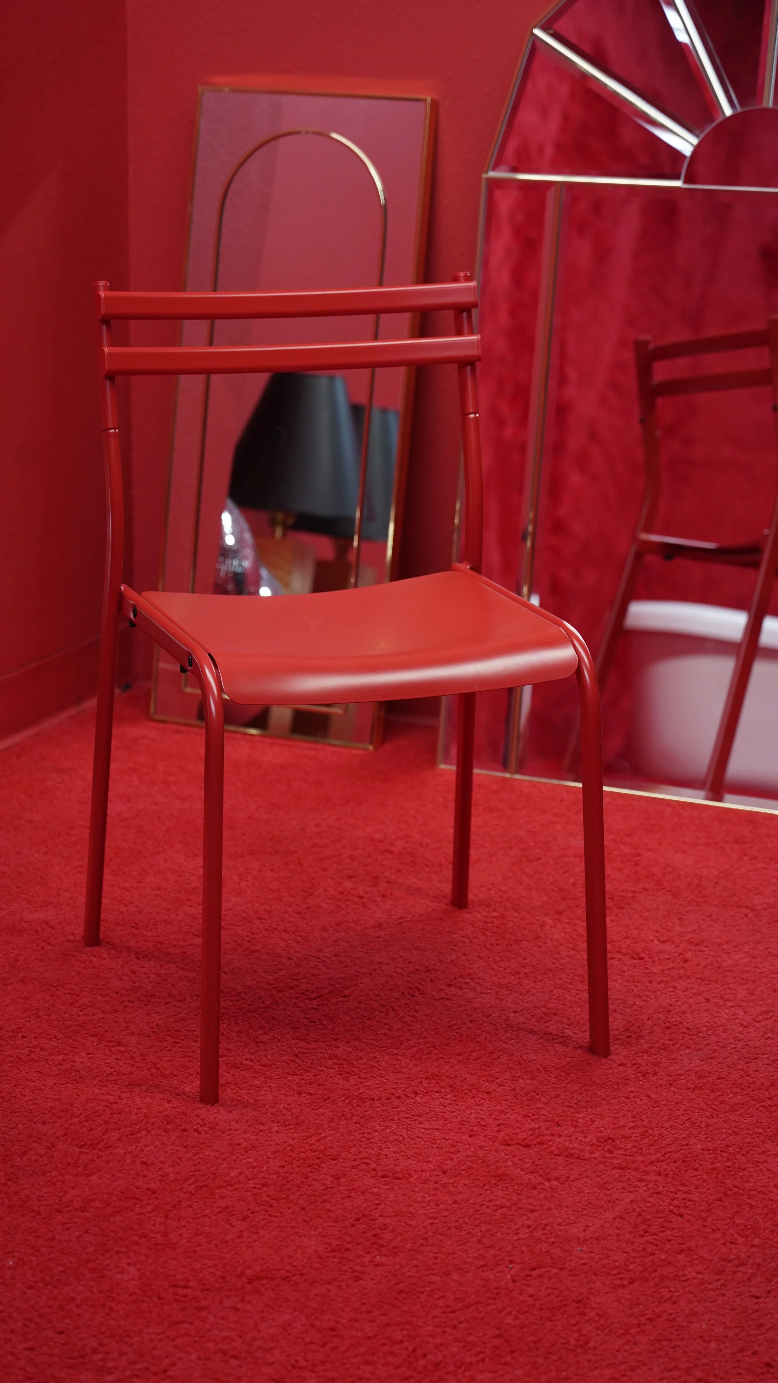 A red chair with a metal frame is positioned in front of a mirror reflecting a black lamp and some decor. The room has red walls and red carpet, with additional red furniture and a decorative mirror visible.
