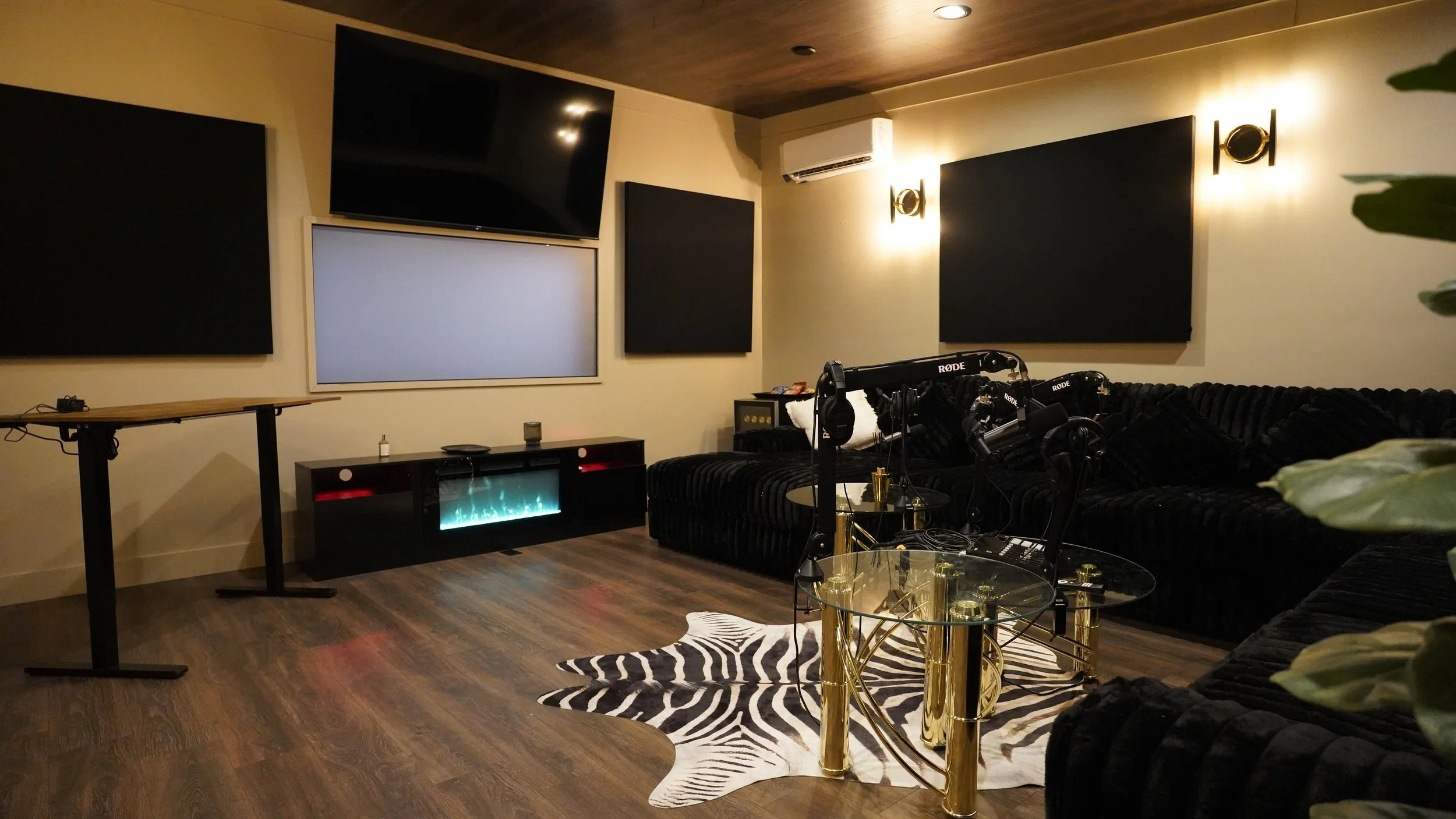 A modern home theater room with large black soundproof panels on the beige walls, a flat-screen TV mounted high on the wall, and a black cozy sectional sofa. There is a glass coffee table with gold accents, a zebra-patterned rug, and a small black en