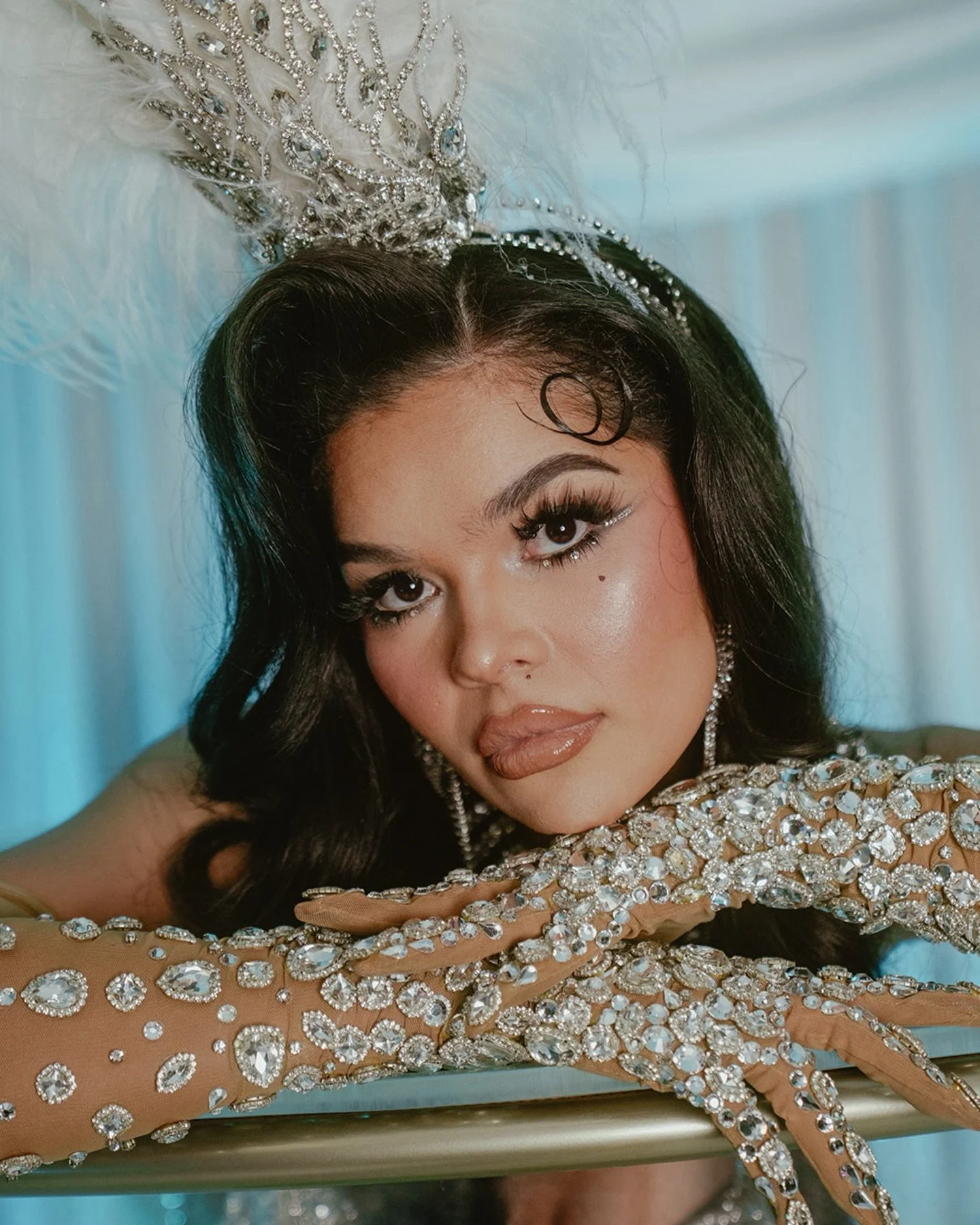 A woman with long dark hair and makeup posing with her face resting on her hand. She is wearing a sparkling, bejeweled dress and a large tiara with feathers on her head.