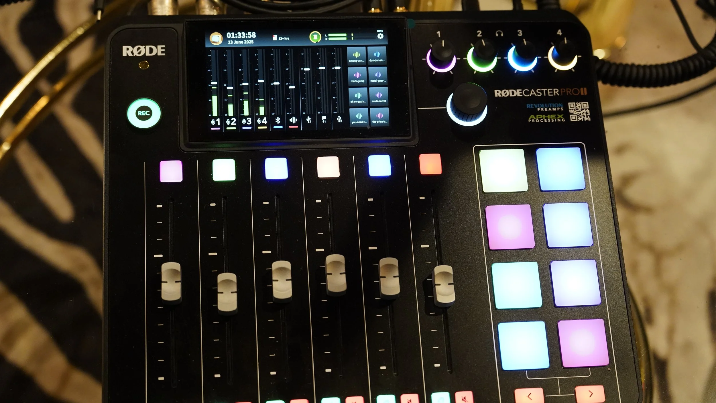 A digital audio mixer with a small touchscreen display, colored buttons, and sliders, labeled Rodecaster Pro II, used for audio production.
