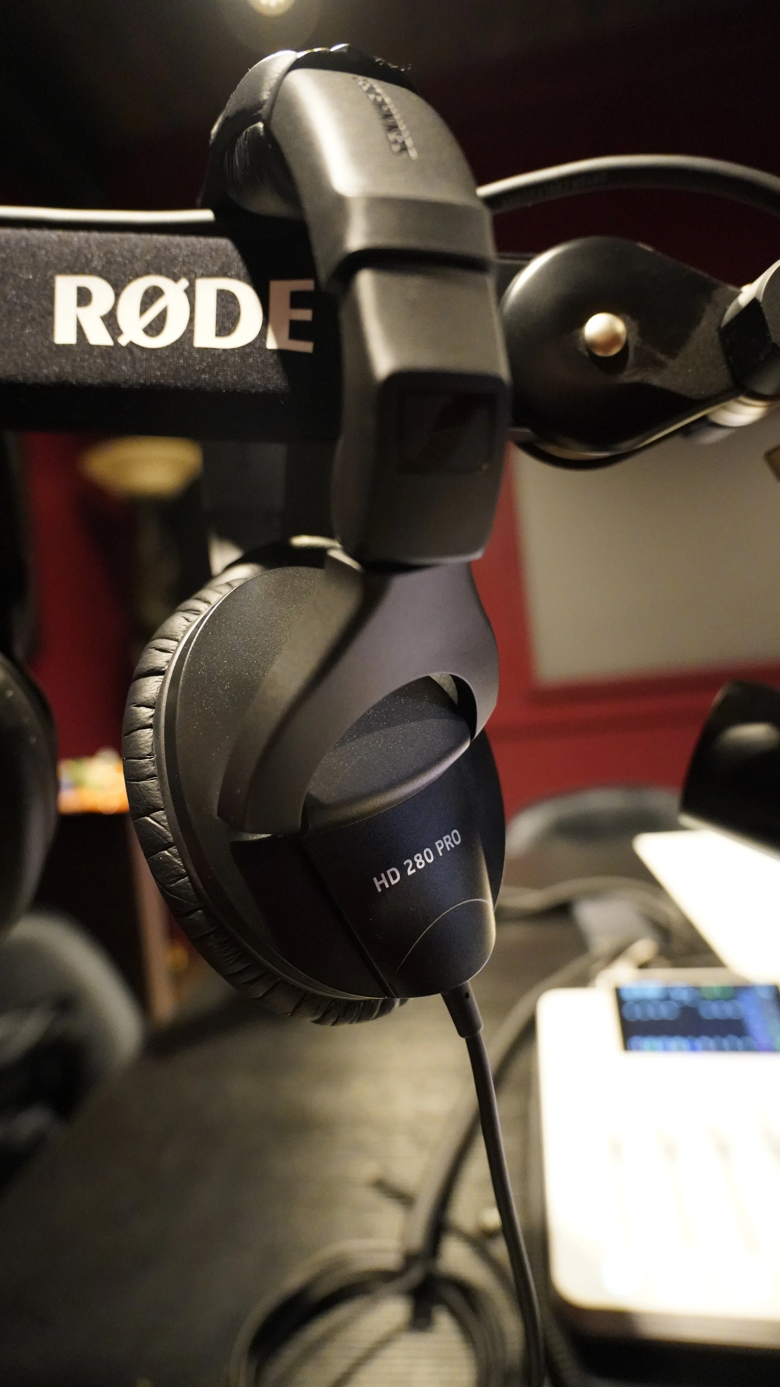 A close-up view of a professional headset labeled HD 280 PRO hanging on a stand, with a RØDE microphone mounted next to it, in a recording or editing studio setting.