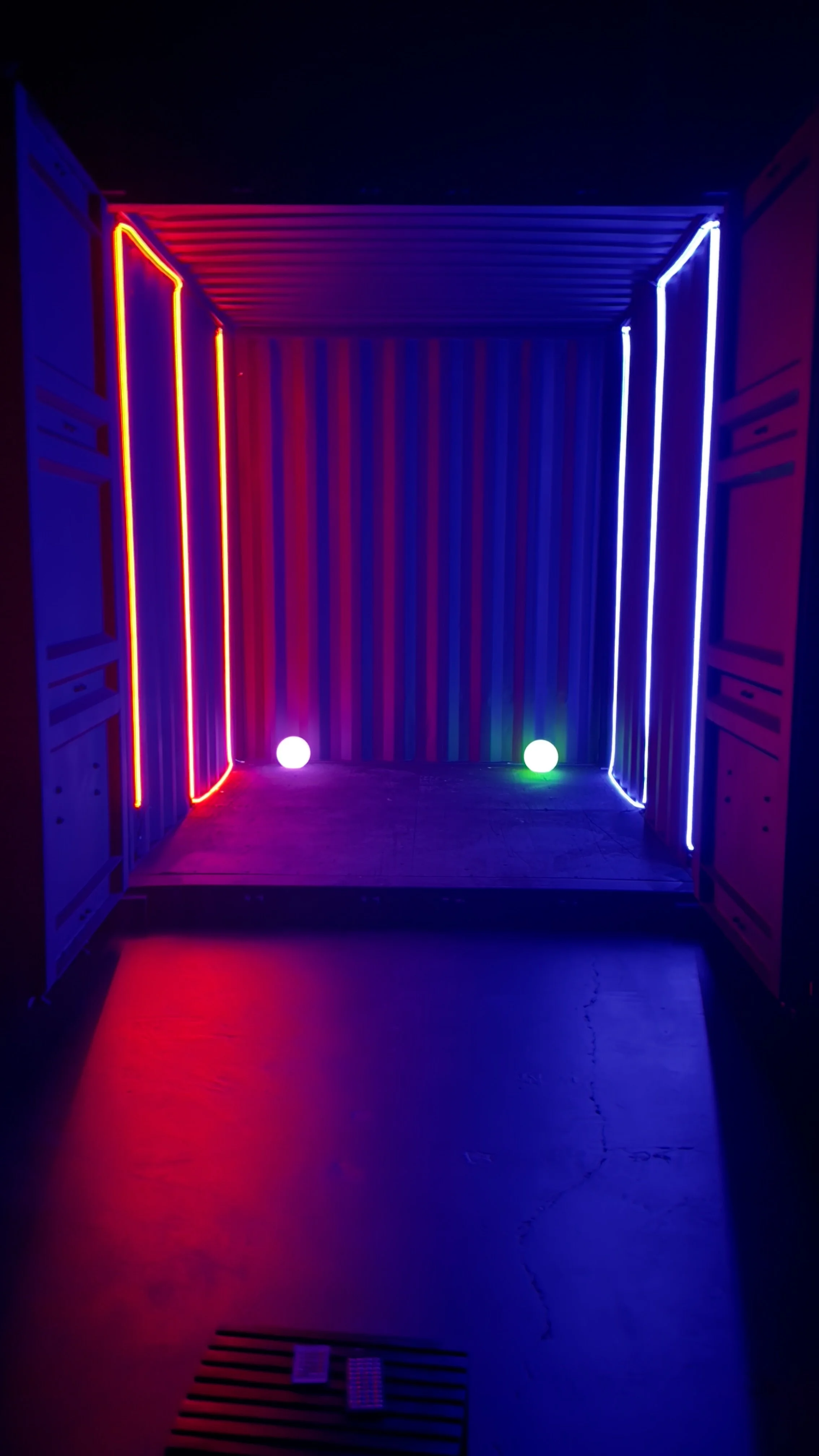 Dark room with colorful neon lights on the walls, two glowing spheres on the floor, and striped background with a stage.
