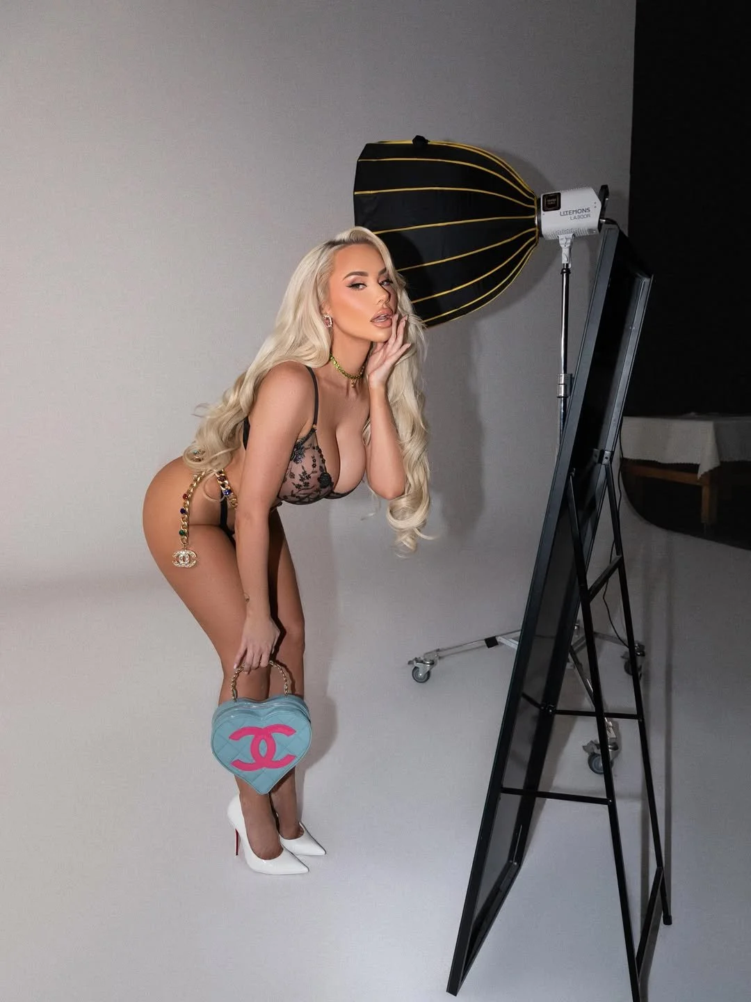 A woman with long blonde hair poses in a photography studio, wearing lingerie, white high heels, and holding a blue heart-shaped Chanel bag. She is standing on a white backdrop with a large studio light and reflector nearby.
