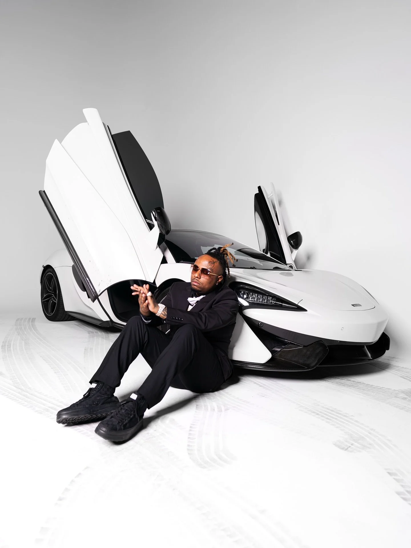 A man with dreadlocks, sunglasses, and black clothing sitting on the floor in front of a white sports car with scissor doors, posing confidently.