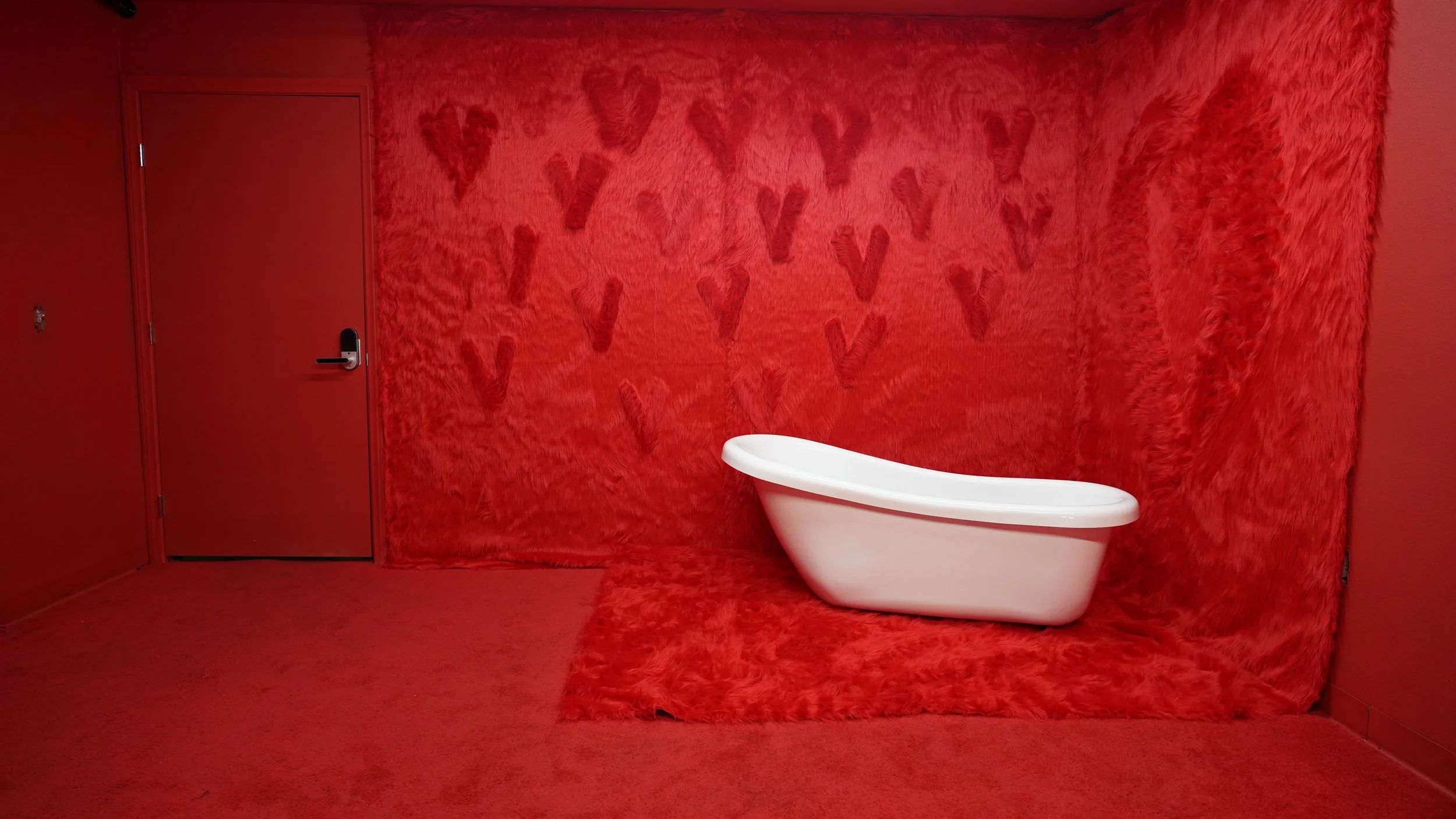 Room with red faux fur walls, floor, and ceiling, decorated with red heart shapes on the back wall. A white bathtub is placed on the faux fur on the floor, and there is a closed red door with a black handle.
